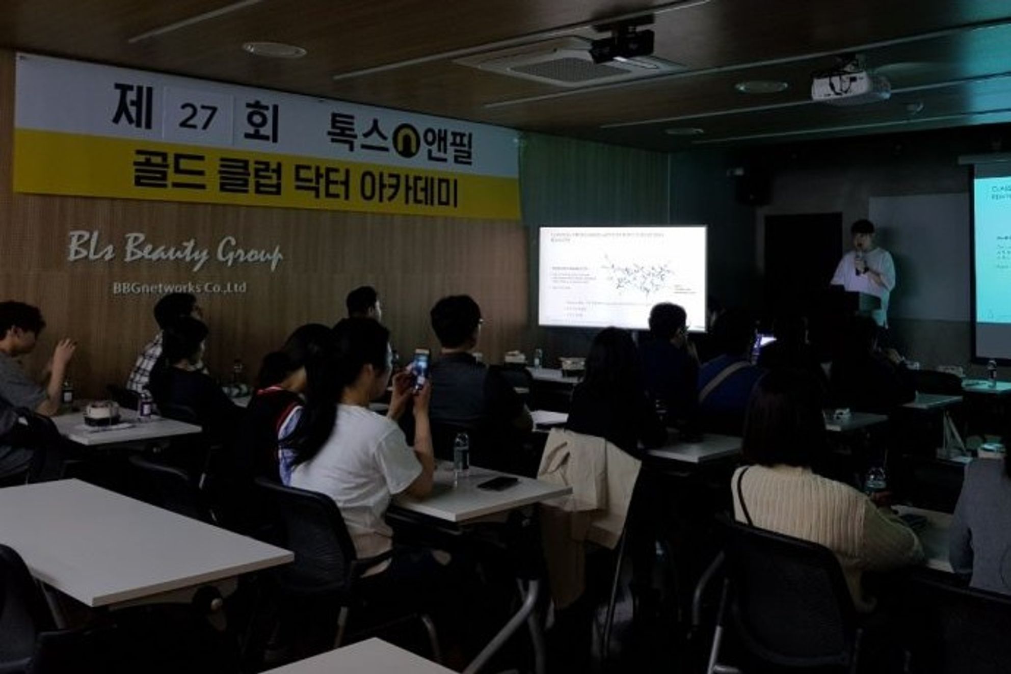 세미나실 앞쪽에서 한 사람이 발표하고 참석자들이 자리에 앉아 있는 장면