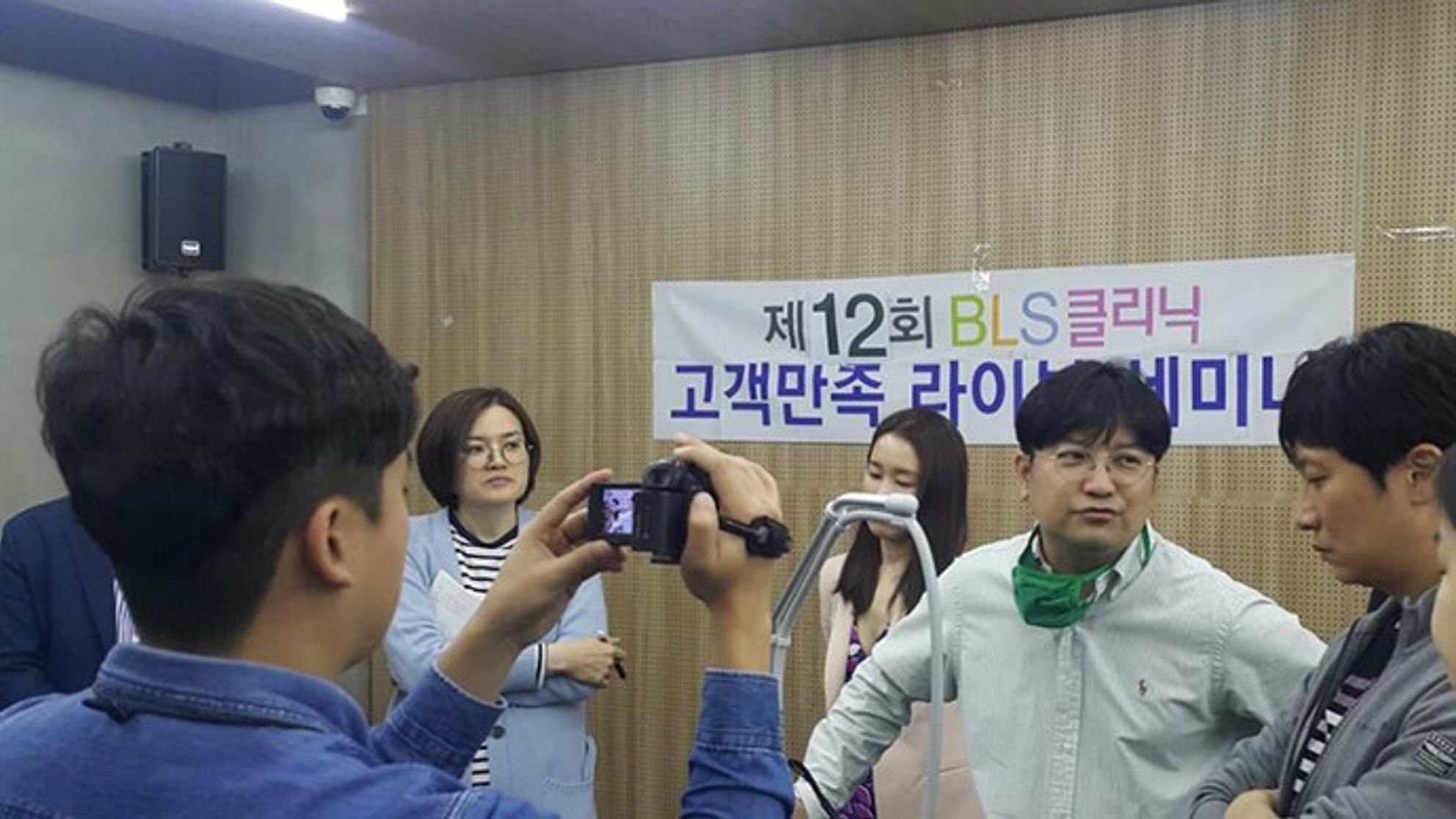 라이브세미나 현수막이 걸린 방에서 여러 사람이 장비 거치대 주위에 서 있고, 앞쪽의 한 사람이 카메라로 장면을 찍는 모습