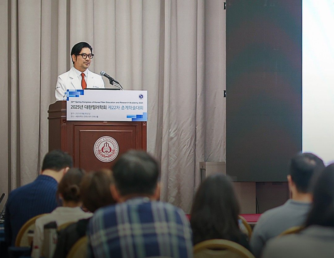 학술대회 이름이 적힌 강연대에서 흰 가운 차림의 한 사람이 말하고, 앞쪽에 청중의 뒷모습이 보이는 모습