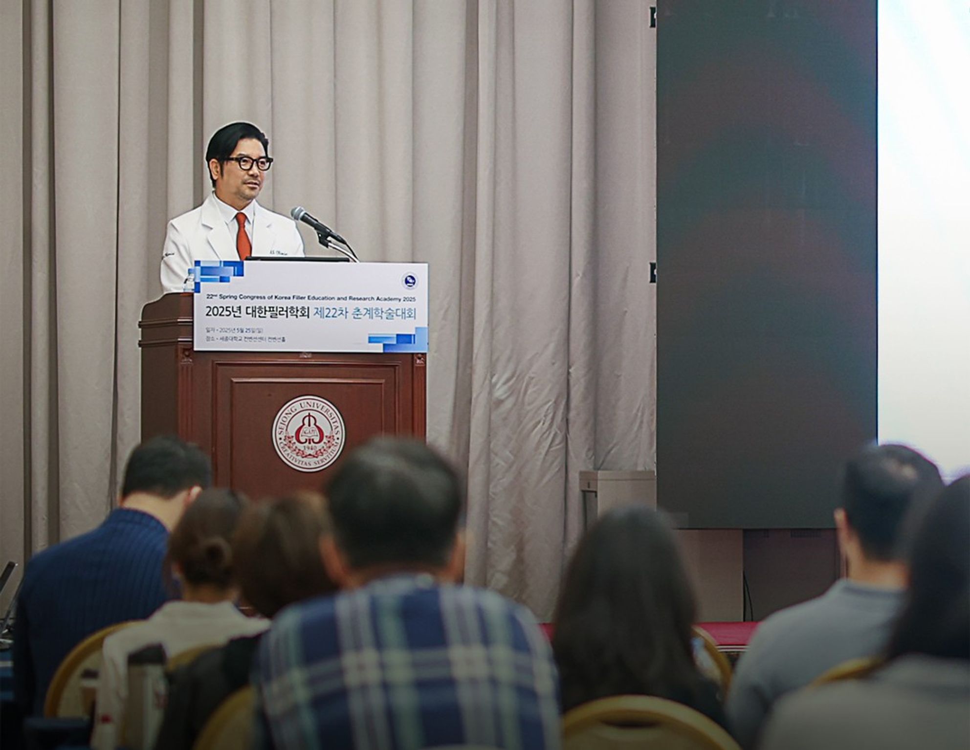 학술대회 이름이 적힌 강연대에서 흰 가운 차림의 한 사람이 말하고, 앞쪽에 청중의 뒷모습이 보이는 모습
