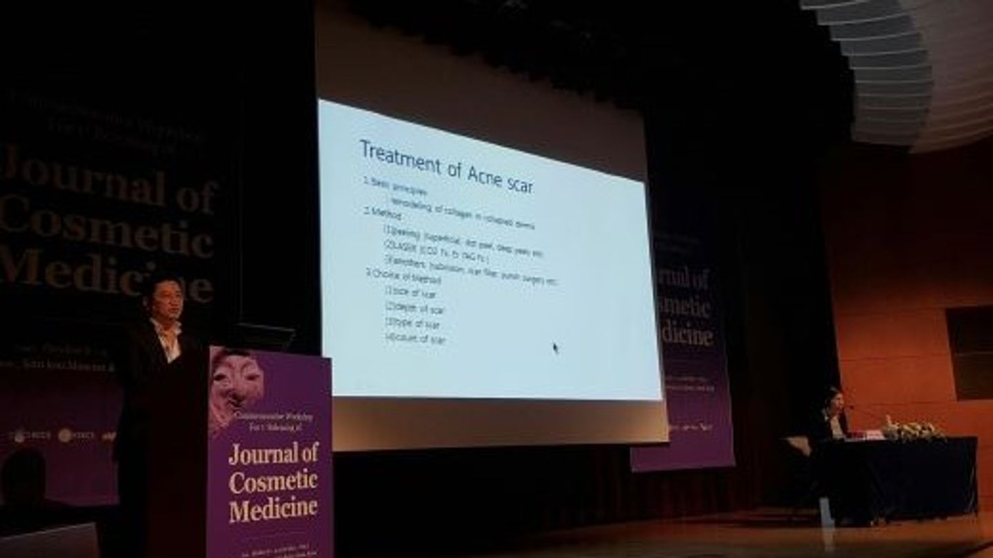 어두운 강당 무대 왼쪽 연단에 한 사람이 서 있고, 가운데 화면에 영문 강의 슬라이드가 떠 있으며, 오른쪽 탁자에 한 사람이 앉아 있는 모습