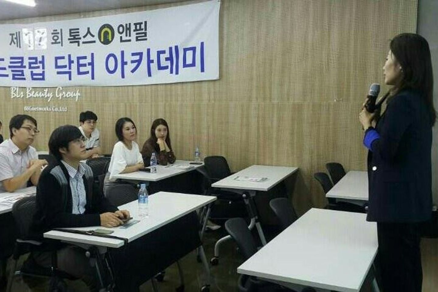 오른쪽에 선 사람이 마이크를 들고 말하고, 왼쪽 책상에 앉은 참석자들이 그쪽을 바라보는 모습
