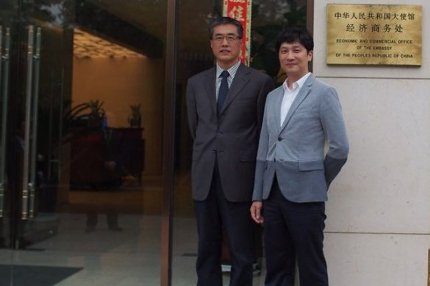 중화인민공화국 대사관 경제상무처 현판이 걸린 건물 유리문 입구 옆에 정장 차림의 두 사람이 나란히 서 있는 모습