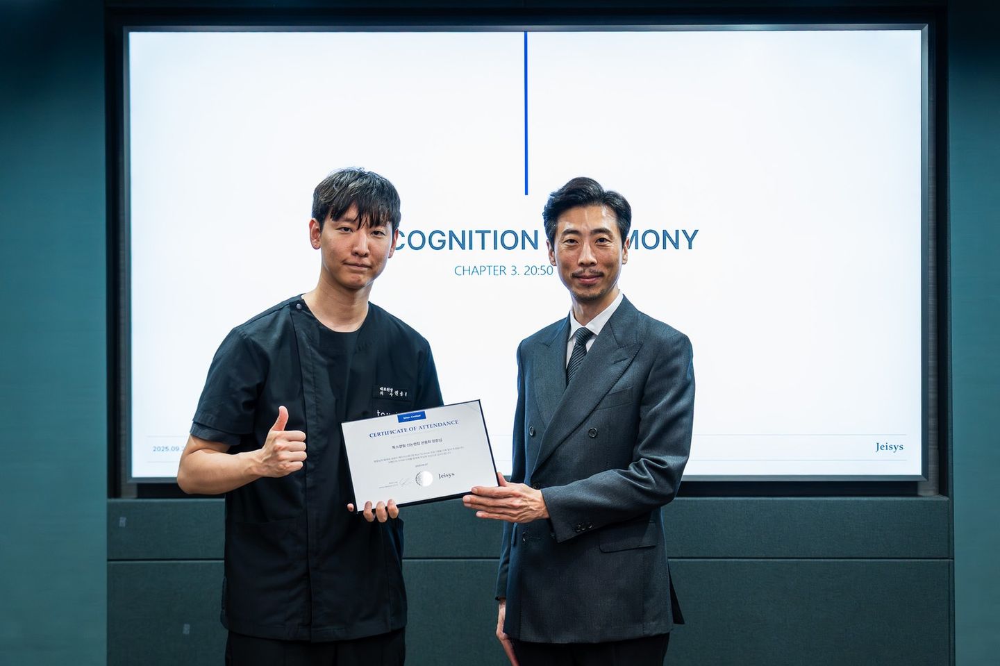 'RECOGNITION CEREMONY' 화면 앞에서 검은 진료복 차림의 남성과 정장 차림의 남성이 'CERTIFICATE OF ATTENDANCE' 증서를 함께 들고 서 있다