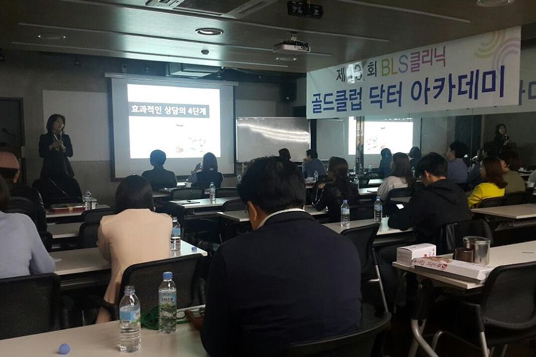 BLS클리닉 현수막이 걸린 강의장에서 참석자들이 책상에 앉아 앞쪽 스크린과 마이크를 든 사람을 바라보는 뒷모습