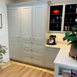 A set of kitchen cabinets with light grey doors and frames sit to the left of a white stone worktop. There is a display cabinet with some glasses and a red crock pot. A large potted plan sits in the bottom left corner.