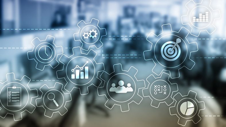 A photo of gears fitting together that depict different tasks a company has to complete in front of a blurred photo of an office.