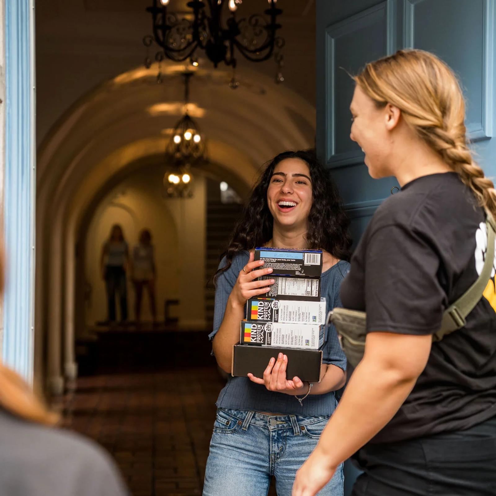 Image of a student laughing while receiving KIND protein bar boxes from a brand ambassador indoors