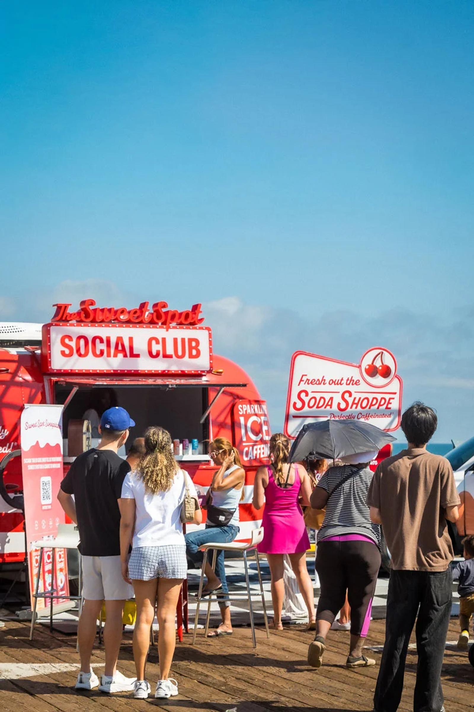 People gather at a red Sparkling Ice “Sweet Spot Social Club” soda shoppe truck during a sunny outdoor event