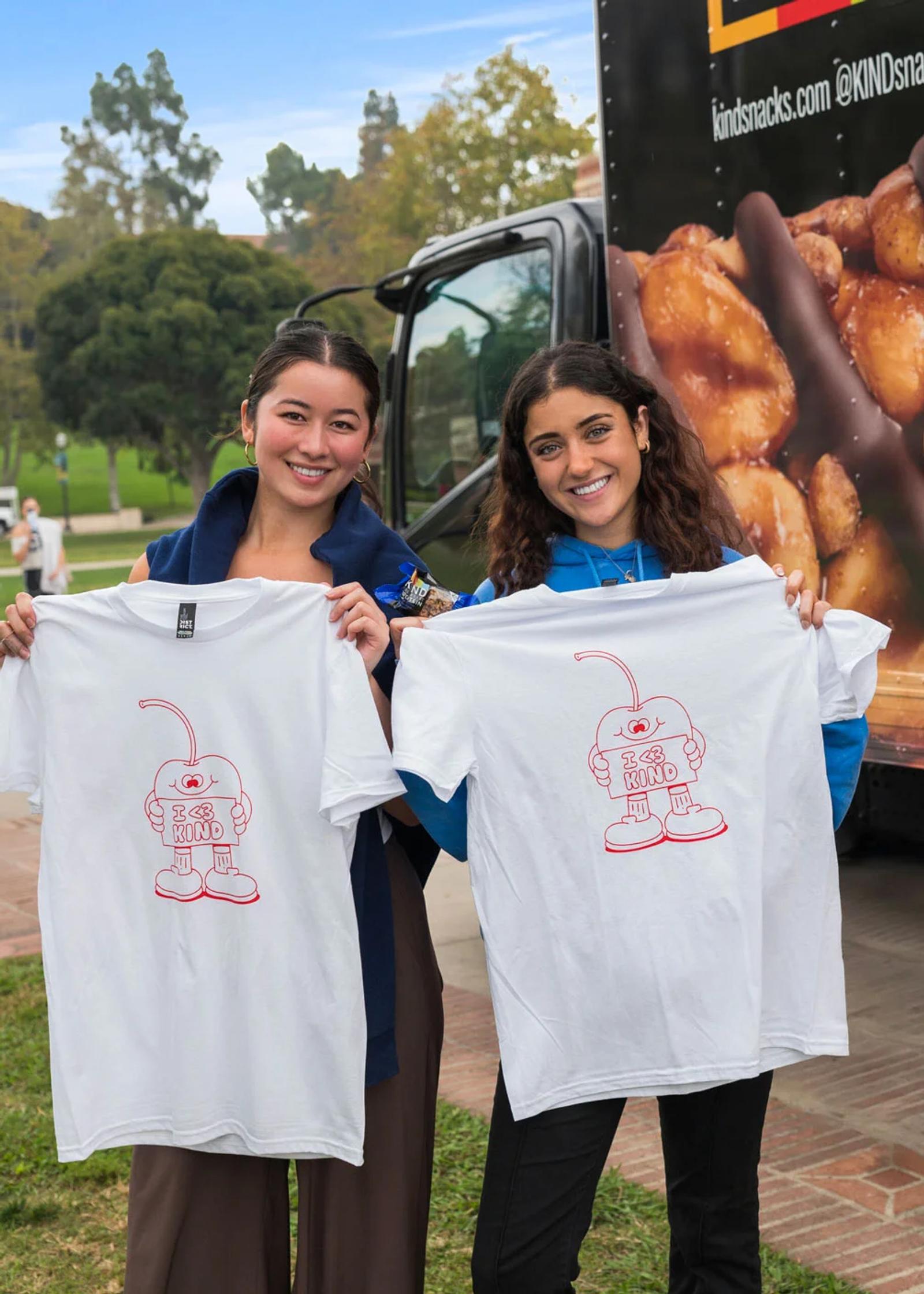 image of two women holding white KIND Snacks branded t-shirts in front of a truck