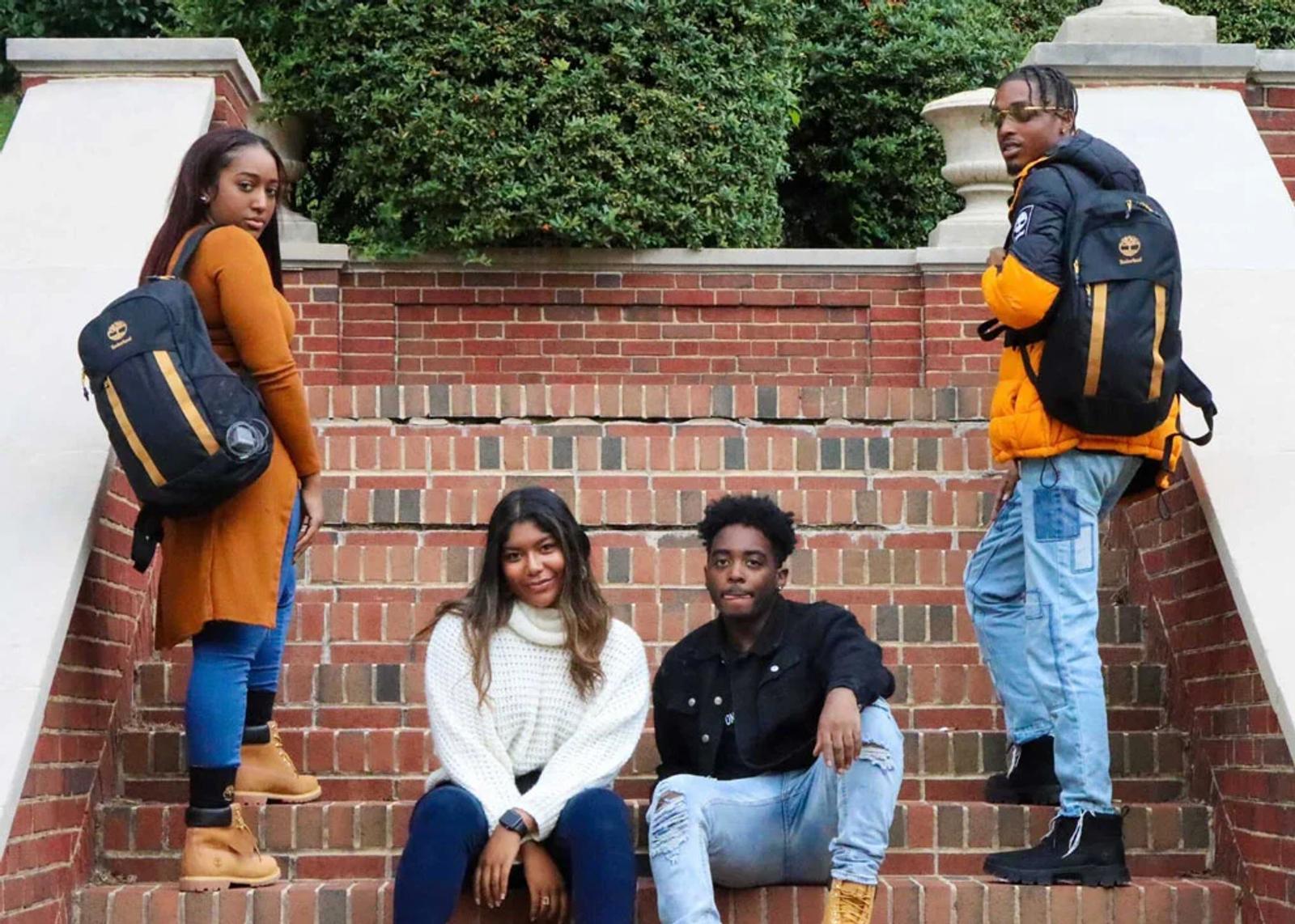 Four young adults wearing Timberland backpacks and boots posed on brick steps outside a campus building