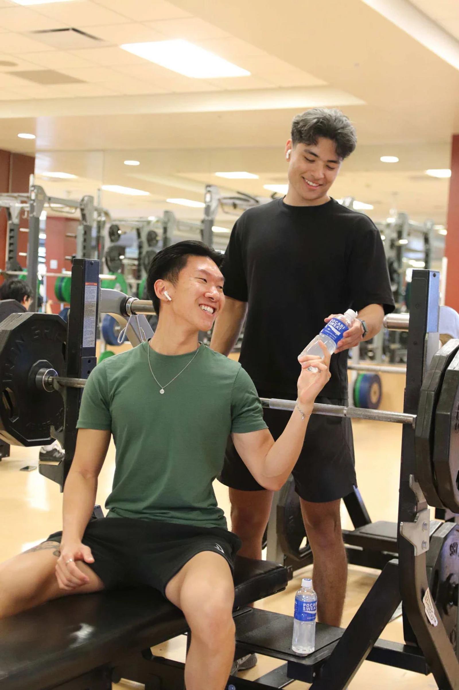 Two young men at the gym, one handing the other a Pocari Sweat bottle between sets