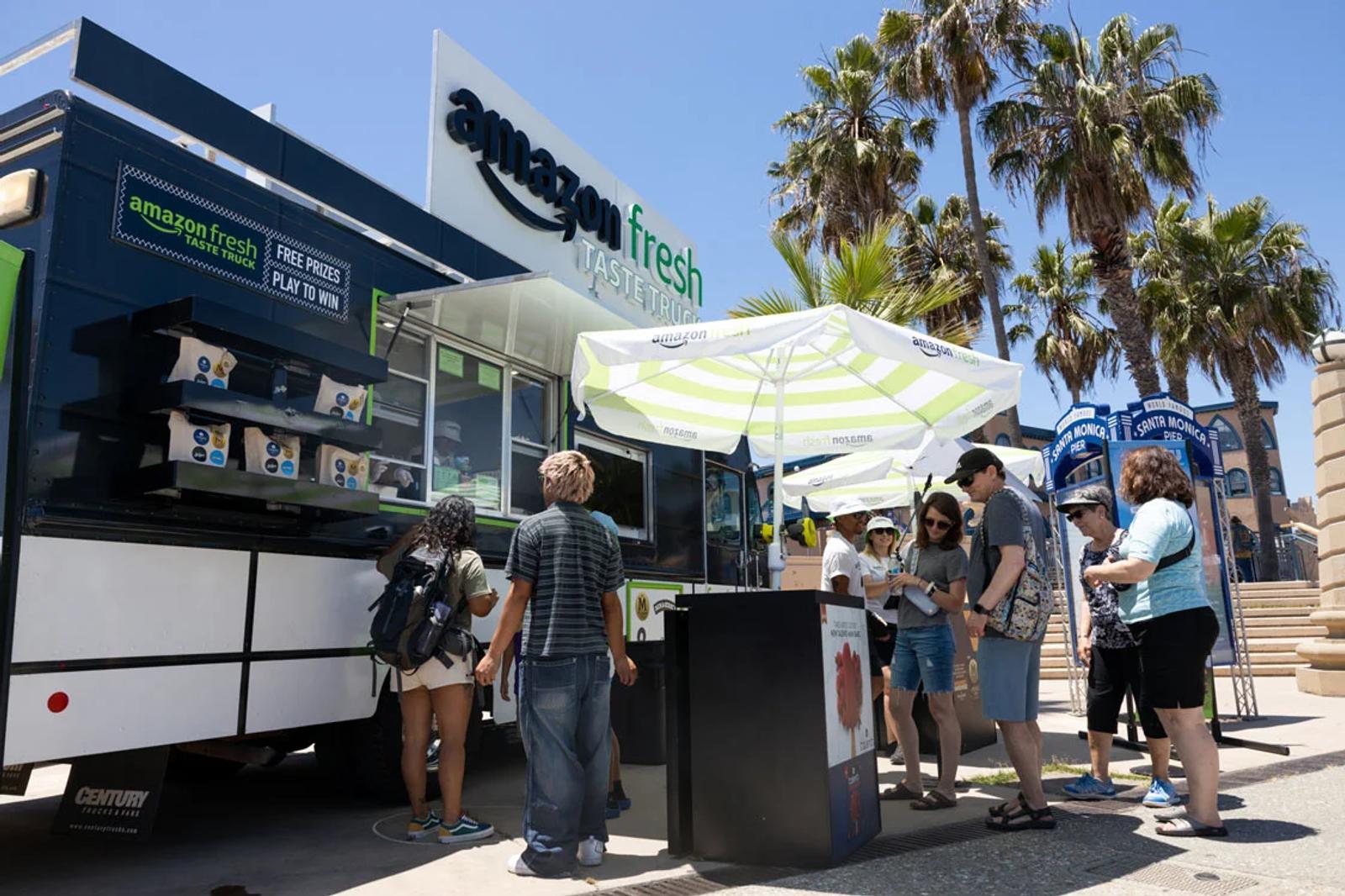 Customers gather around an Amazon Fresh food truck under umbrellas on a sunny day with palm trees in the background