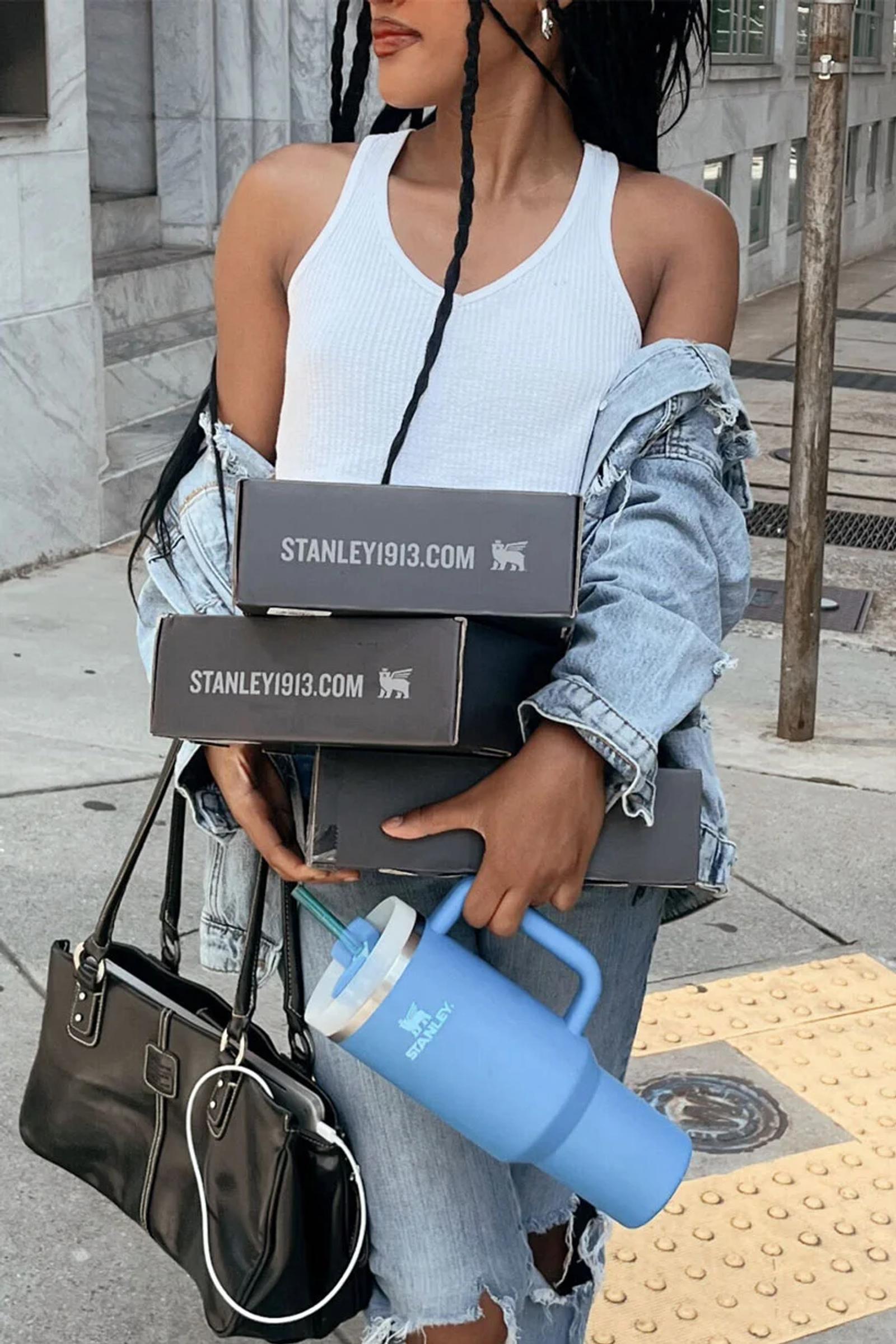 Woman carrying stacked Stanley 1913 product boxes and a blue Stanley tumbler on a city sidewalk