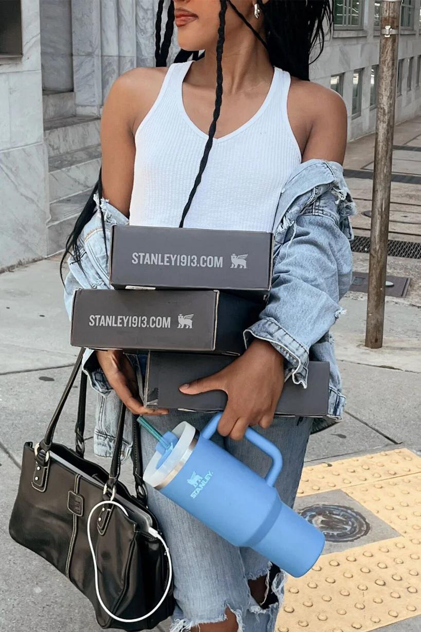 Woman carrying stacked Stanley 1913 product boxes and a blue Stanley tumbler on a city sidewalk