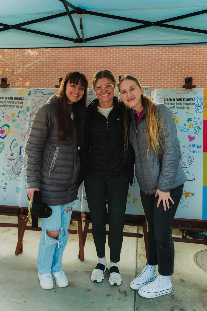Image of three women smiling in front of Color in Some Kindness coloring boards under an event tent