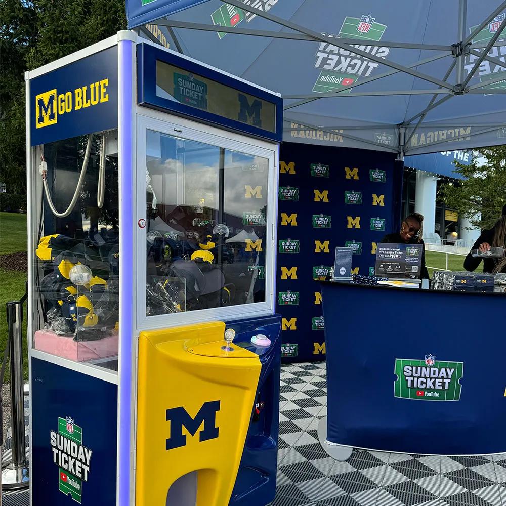 Image of a Michigan-branded claw machine and staffed counter at a YouTube NFL Sunday Ticket activation