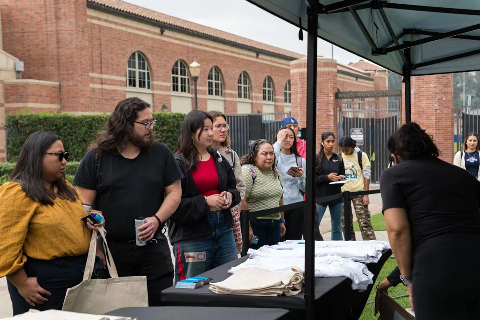 Image of people in line at a KIND activation table with tote bags and T-shirts near UCLA's Wallis Annenberg Stadium