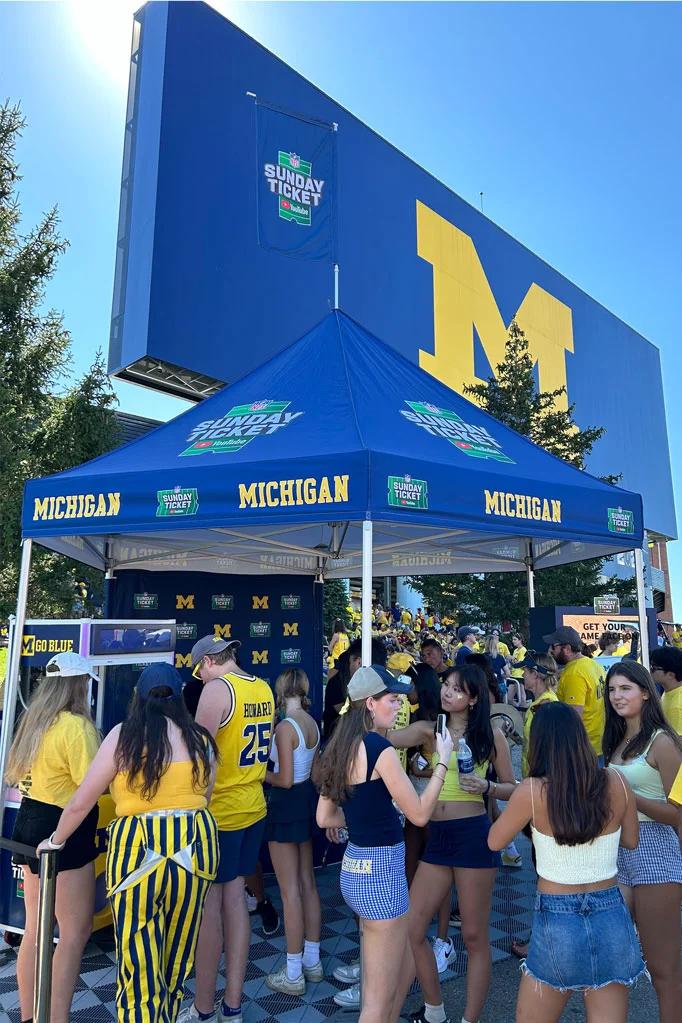 NFL Sunday ticket branded tailgate tent at a university of Michigan football game, with fans in maize and blue