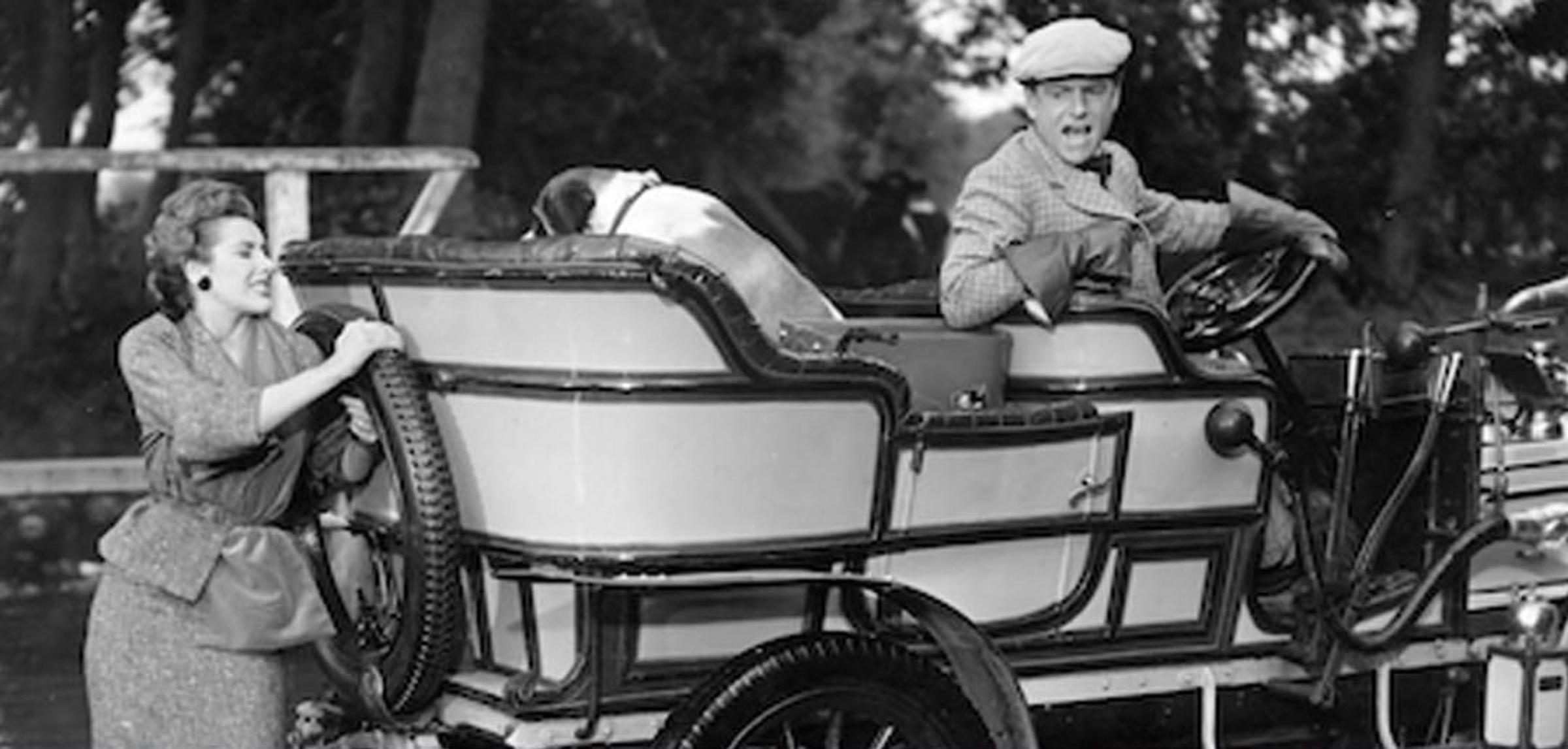 Zwart-wit foto van vrouw die een auto aanduwt en man achter het stuur roept naar haar.