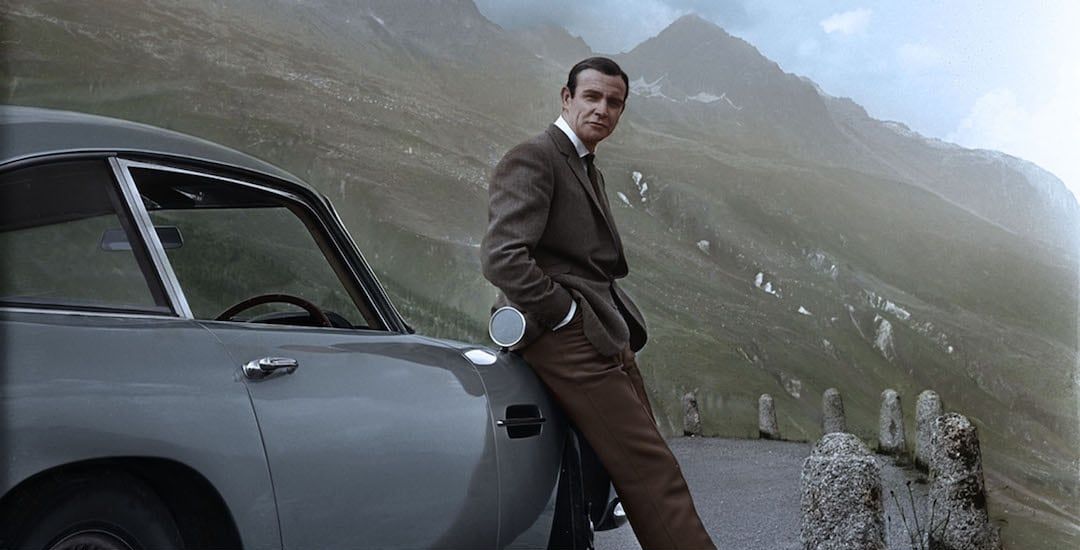 James Bond leunend tegen de Aston Martin DB5 met groene bergen op de achtergrond