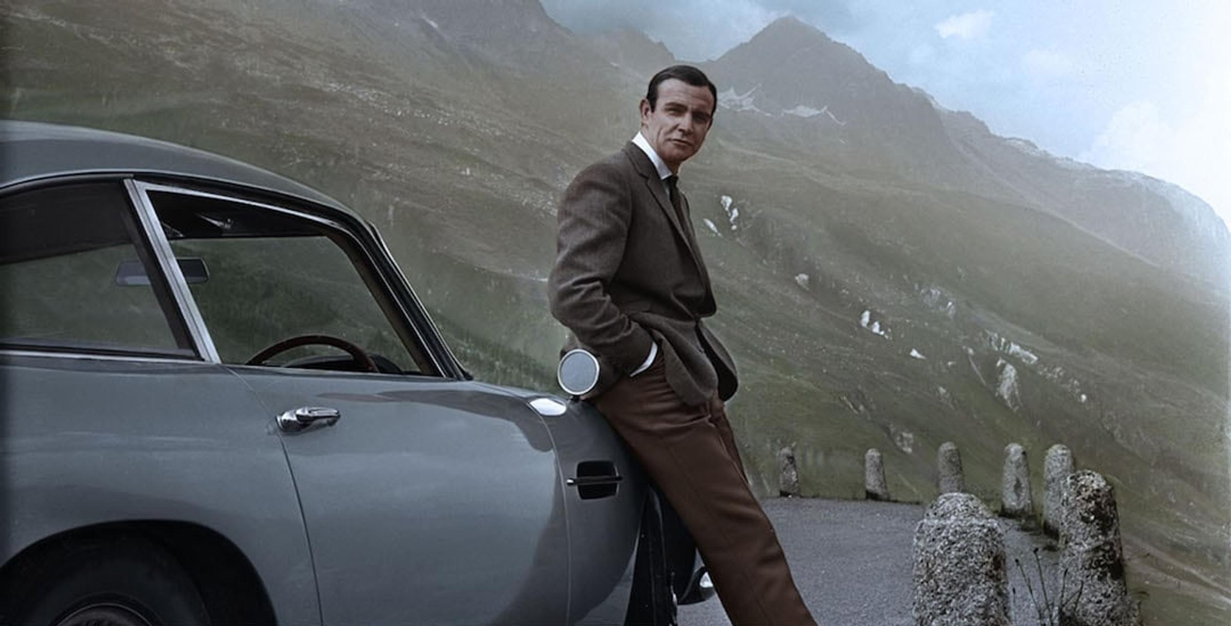 James Bond leunend tegen de Aston Martin DB5 met groene bergen op de achtergrond