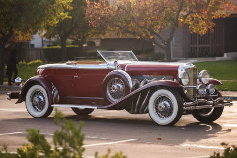 Red Duesenberg car with white wheels and open roof in a car park in a green residential area