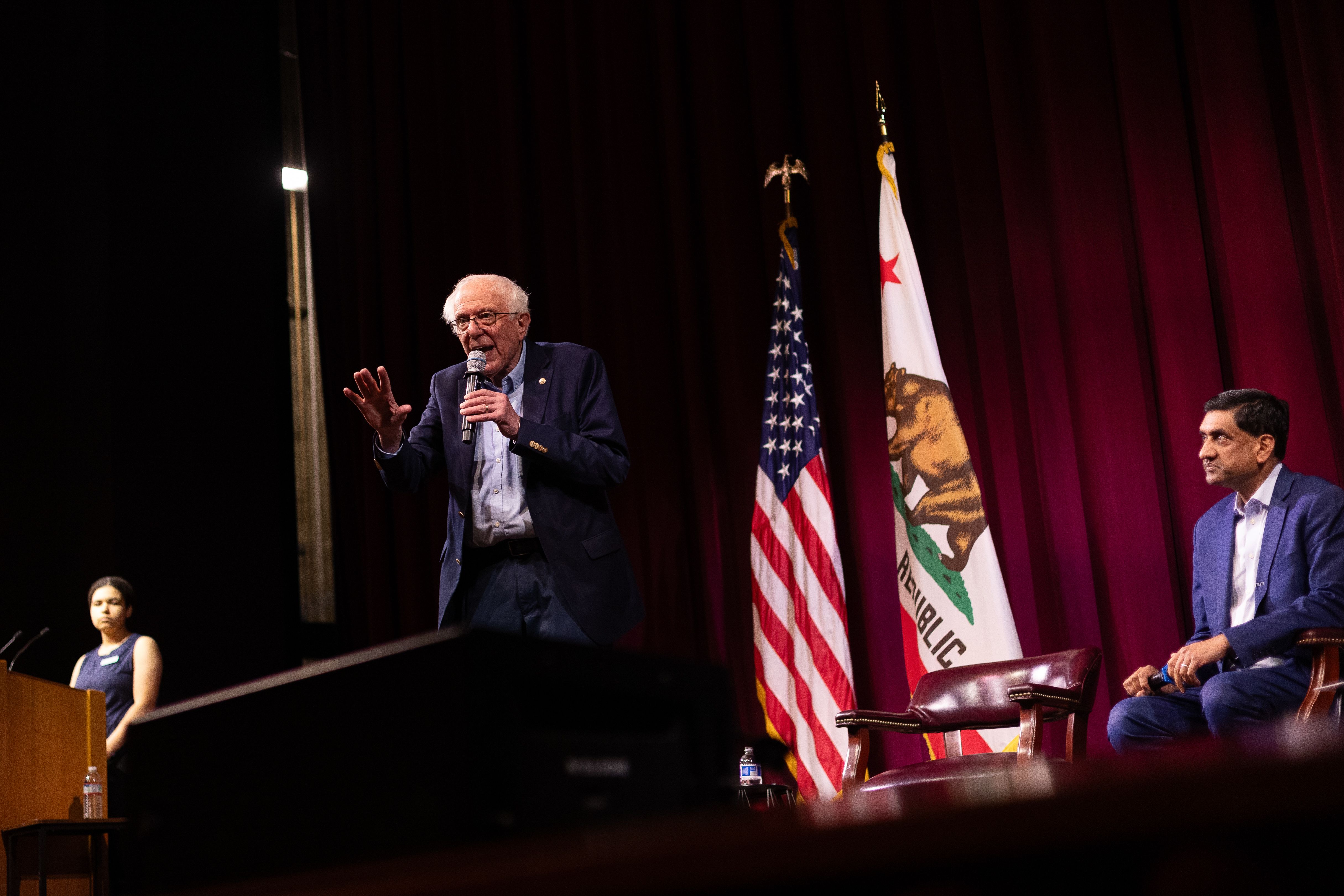 Senator Bernie Sanders stands at a microphone center stage, hand raised mid-gesture, while Representative Ro Khanna sits in a leather chair to the right watching, framed by an American flag and California state flag against a deep red curtain.