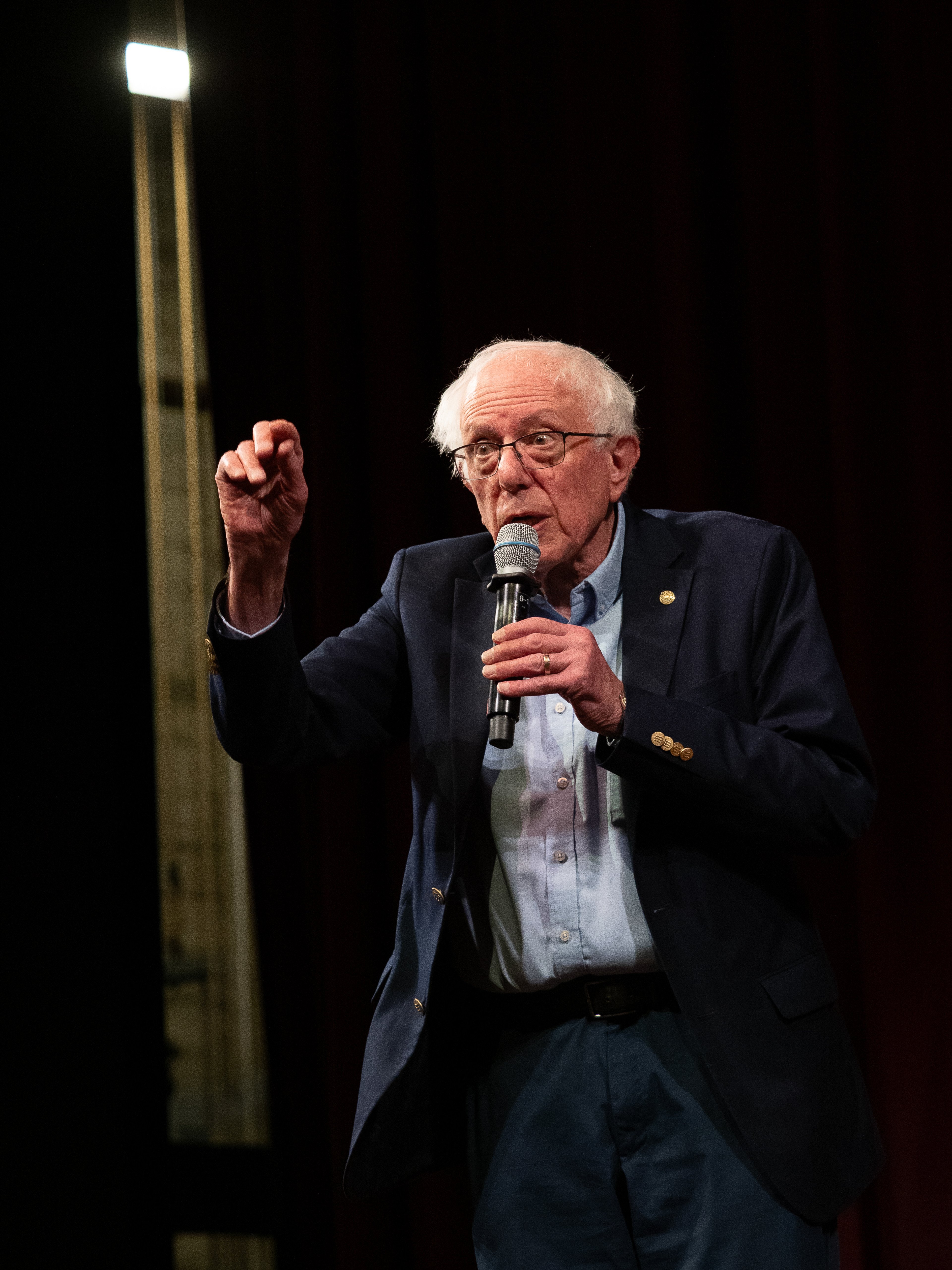 Bernie Sanders standing on stage holding a microphone in one hand, the other raised as a closed fist mid-speech, a stage light visible upper-left against a dark backdrop.