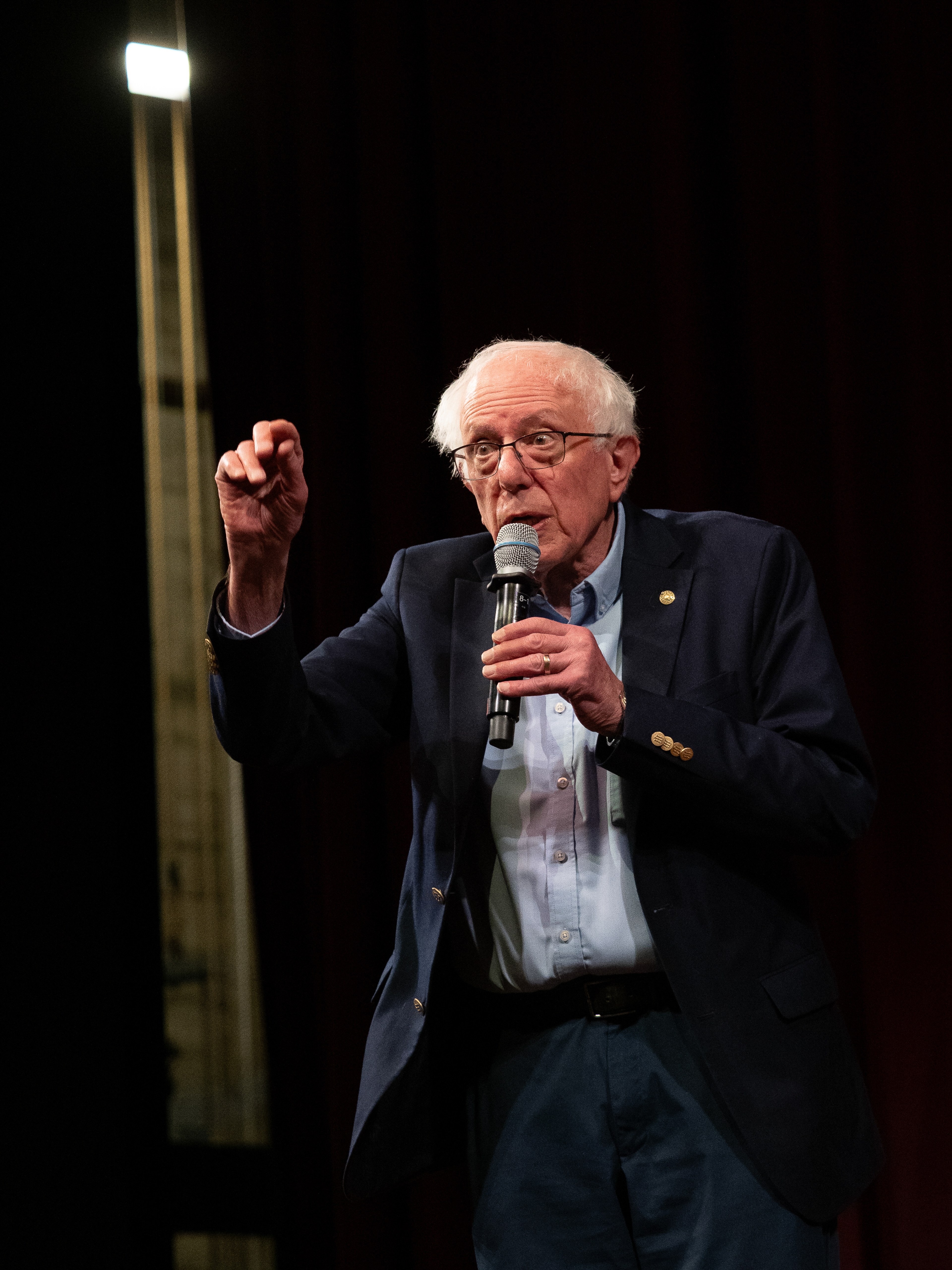 Bernie Sanders standing on stage holding a microphone in one hand, the other raised as a closed fist mid-speech, a stage light visible upper-left against a dark backdrop.
