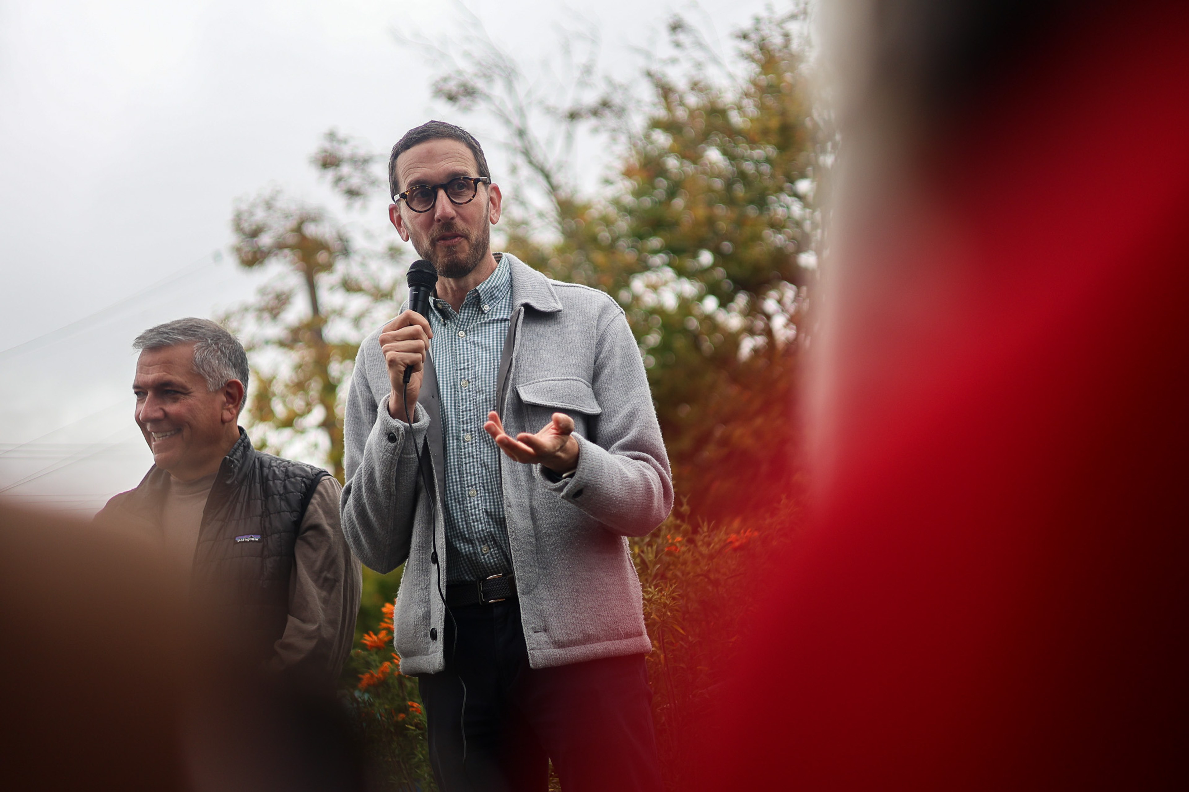 California State Senator Scott Wiener, in glasses and a gray cardigan, speaks into a microphone outdoors with a smiling man in a Patagonia vest beside him, framed loosely by an out-of-focus listener in red.