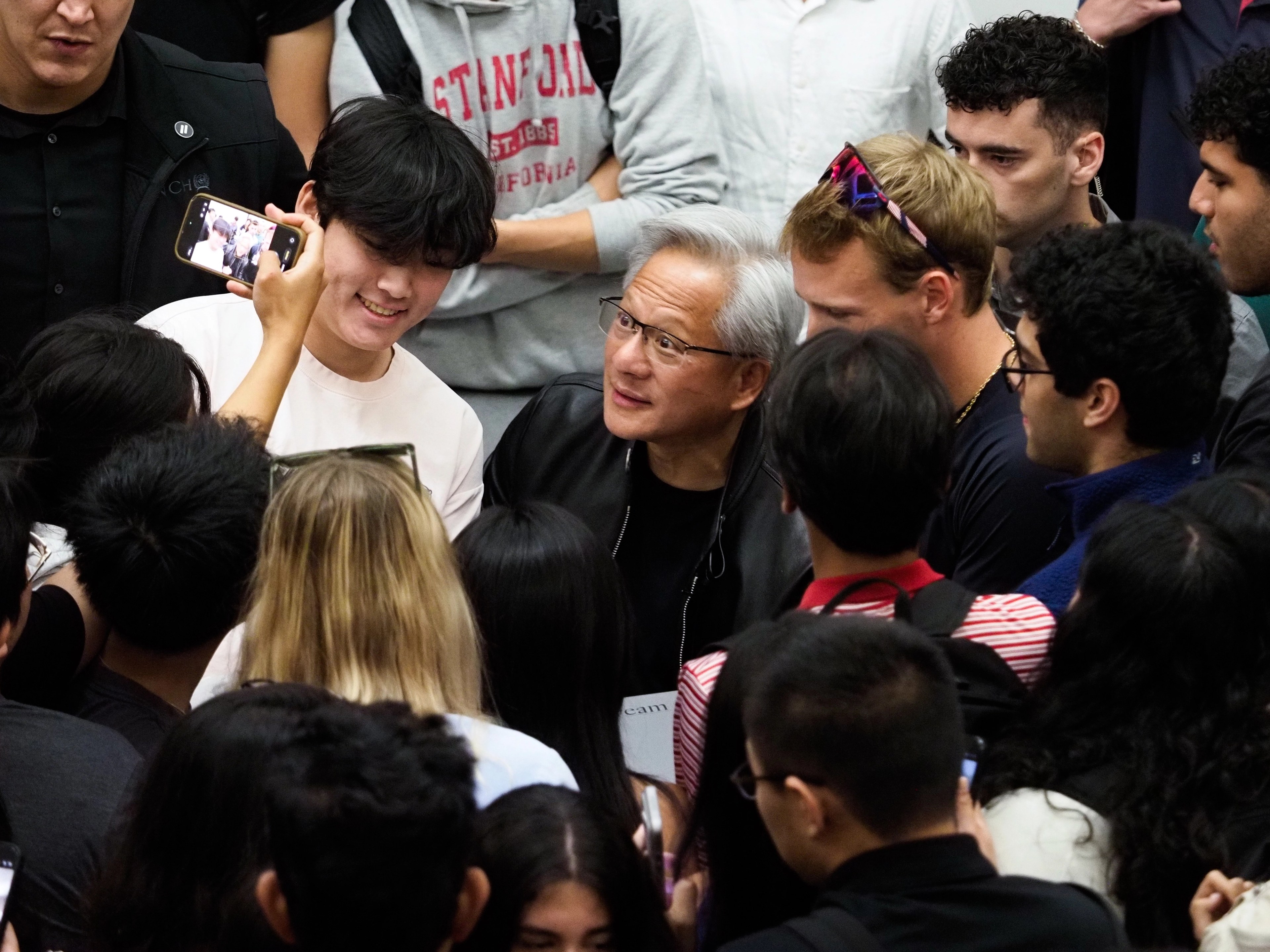 Jensen Huang in the middle of a tight crowd of students, several wearing Stanford sweatshirts, a student in a red Stanford-California sweatshirt visible at upper left, others smiling and pressed in around him.