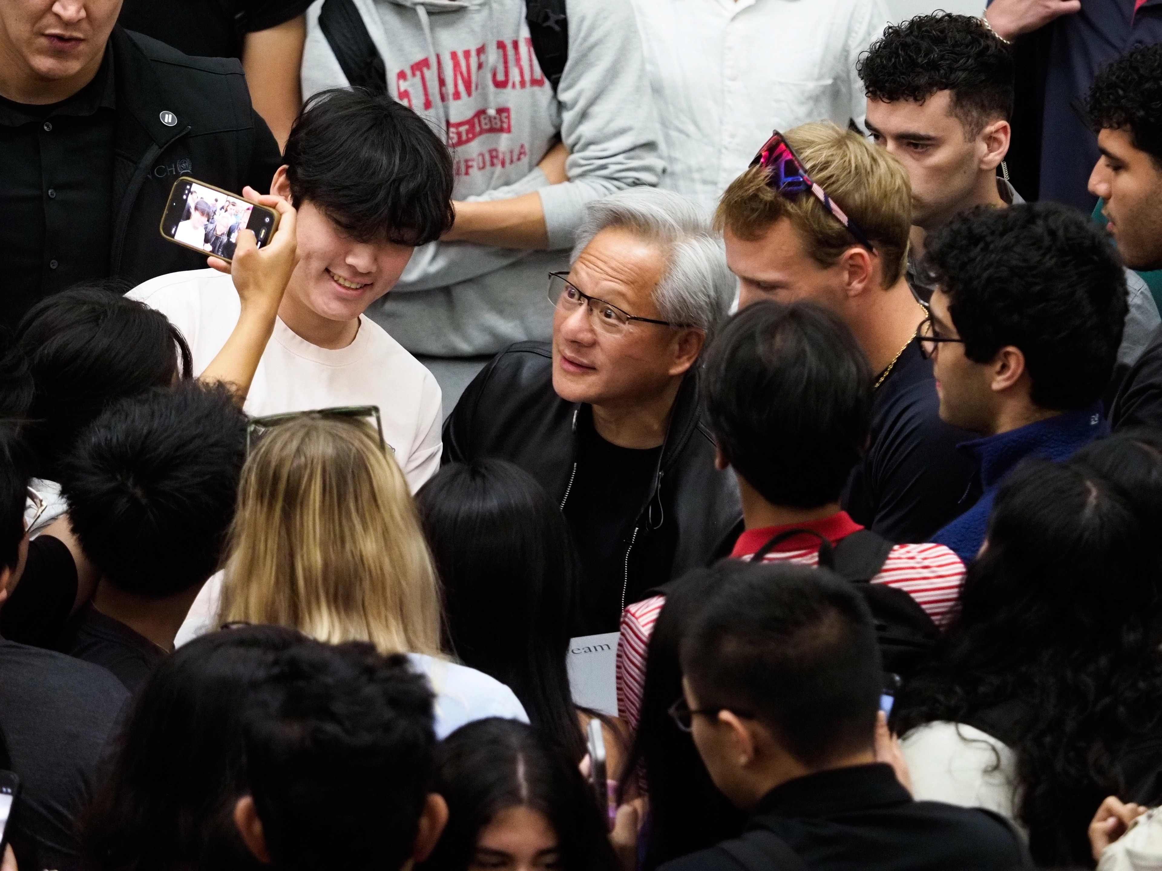 Jensen Huang in the middle of a tight crowd of students, several wearing Stanford sweatshirts, a student in a red Stanford-California sweatshirt visible at upper left, others smiling and pressed in around him.