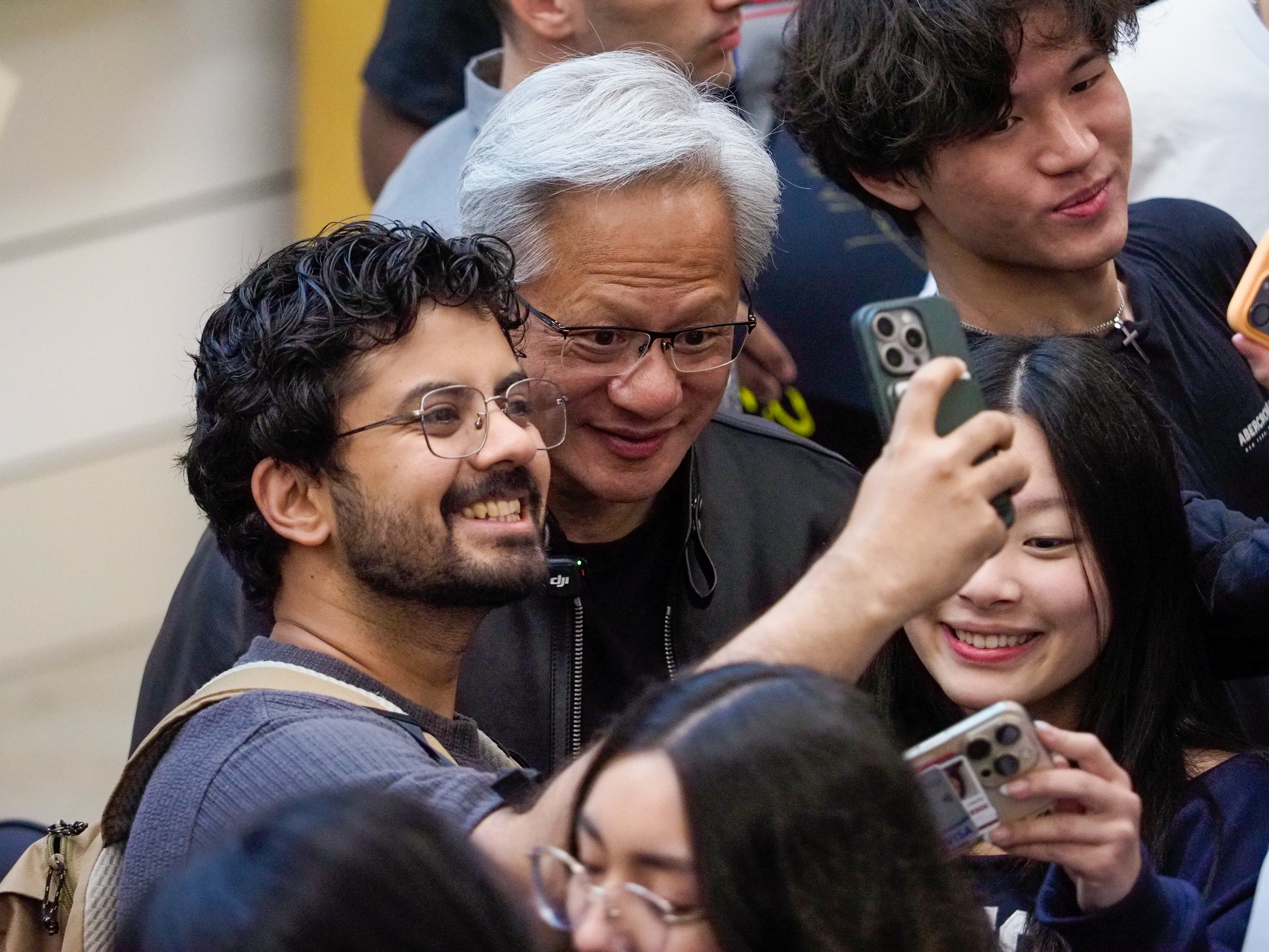 Jensen Huang surrounded by three students taking selfies, his face partially obscured by their phones; a smiling young man in glasses and a young woman in the foreground.