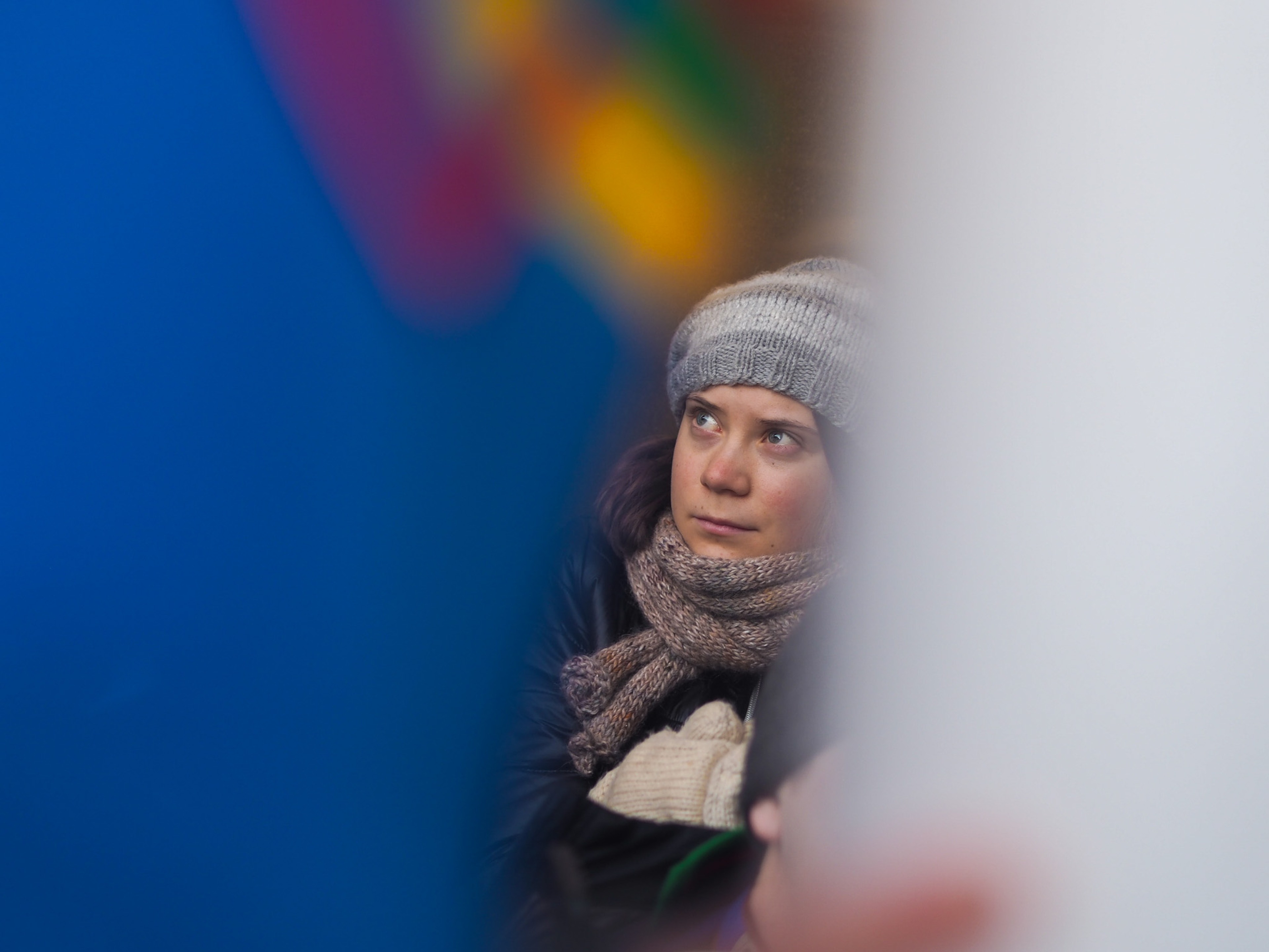 Greta Thunberg in a wool beanie and knit scarf, dark winter coat, photographed through a gap between two out-of-focus protest signs, looking up and to the left.