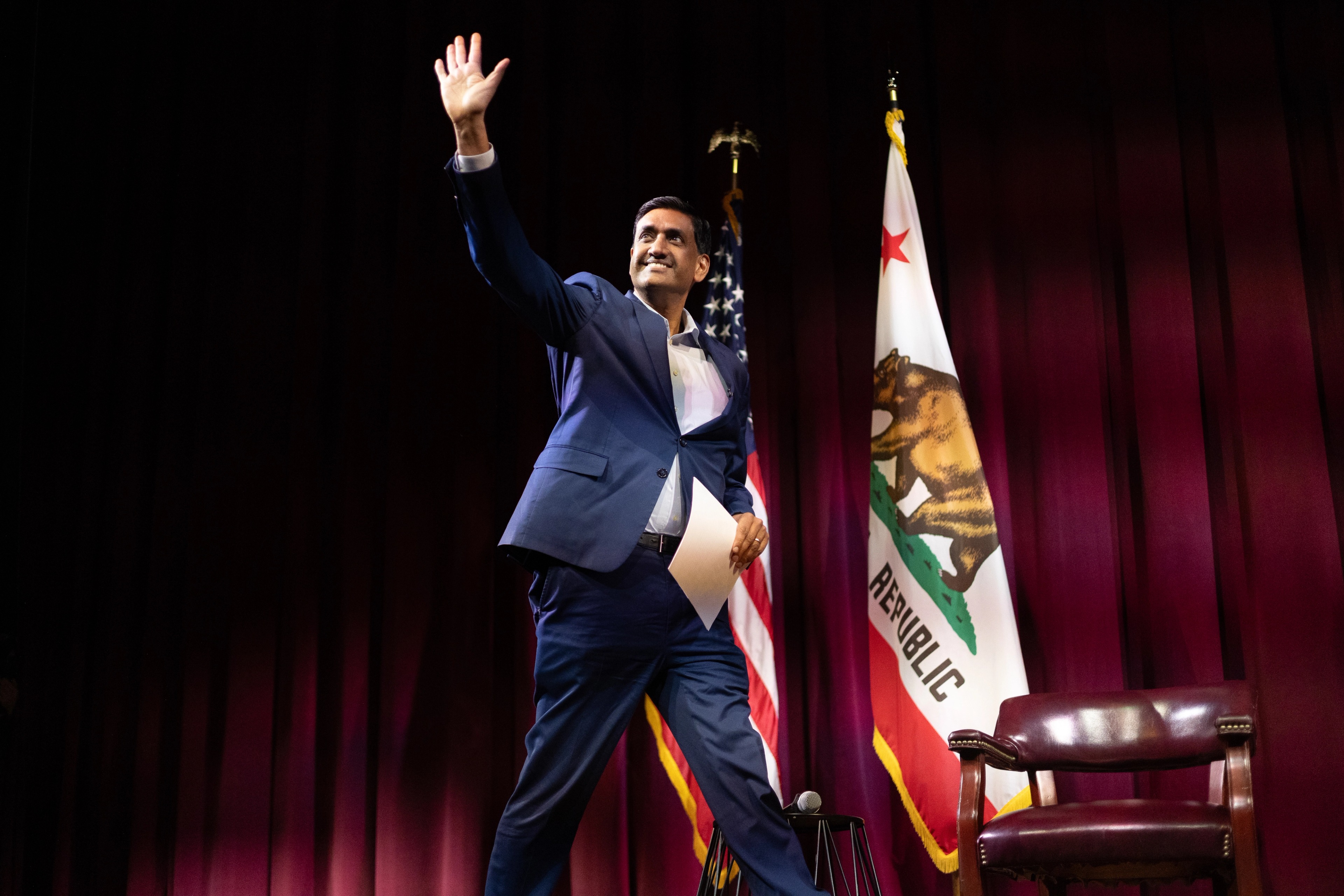 Representative Ro Khanna in a navy suit walks across the stage, arm raised in a wave to the audience, smiling, framed by the American flag and California state flag against a deep red curtain.