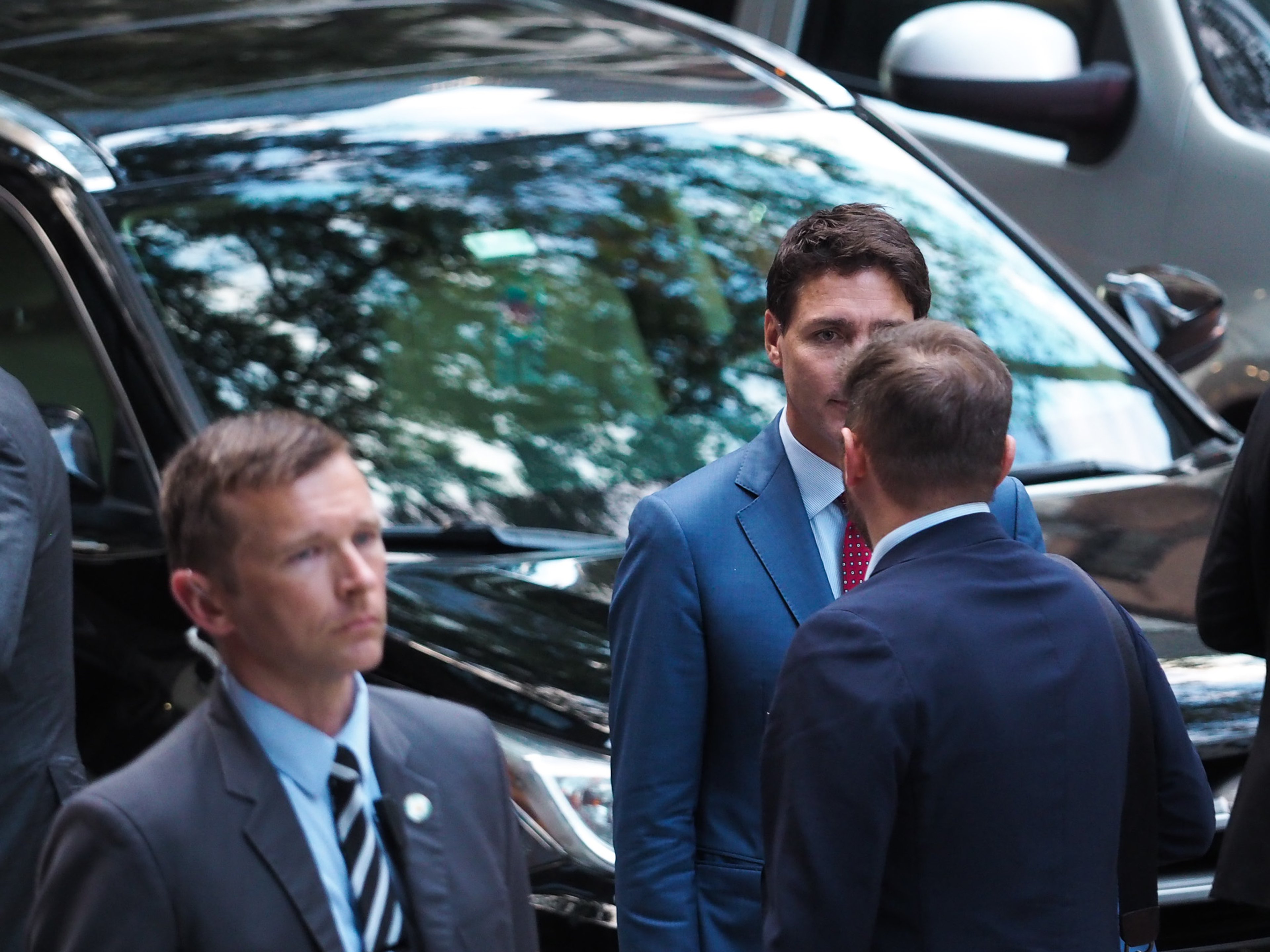 Canadian Prime Minister Justin Trudeau in a blue suit and red dotted tie, mid-conversation between two black motorcade SUVs, an RCMP security agent in a gray suit in the foreground.