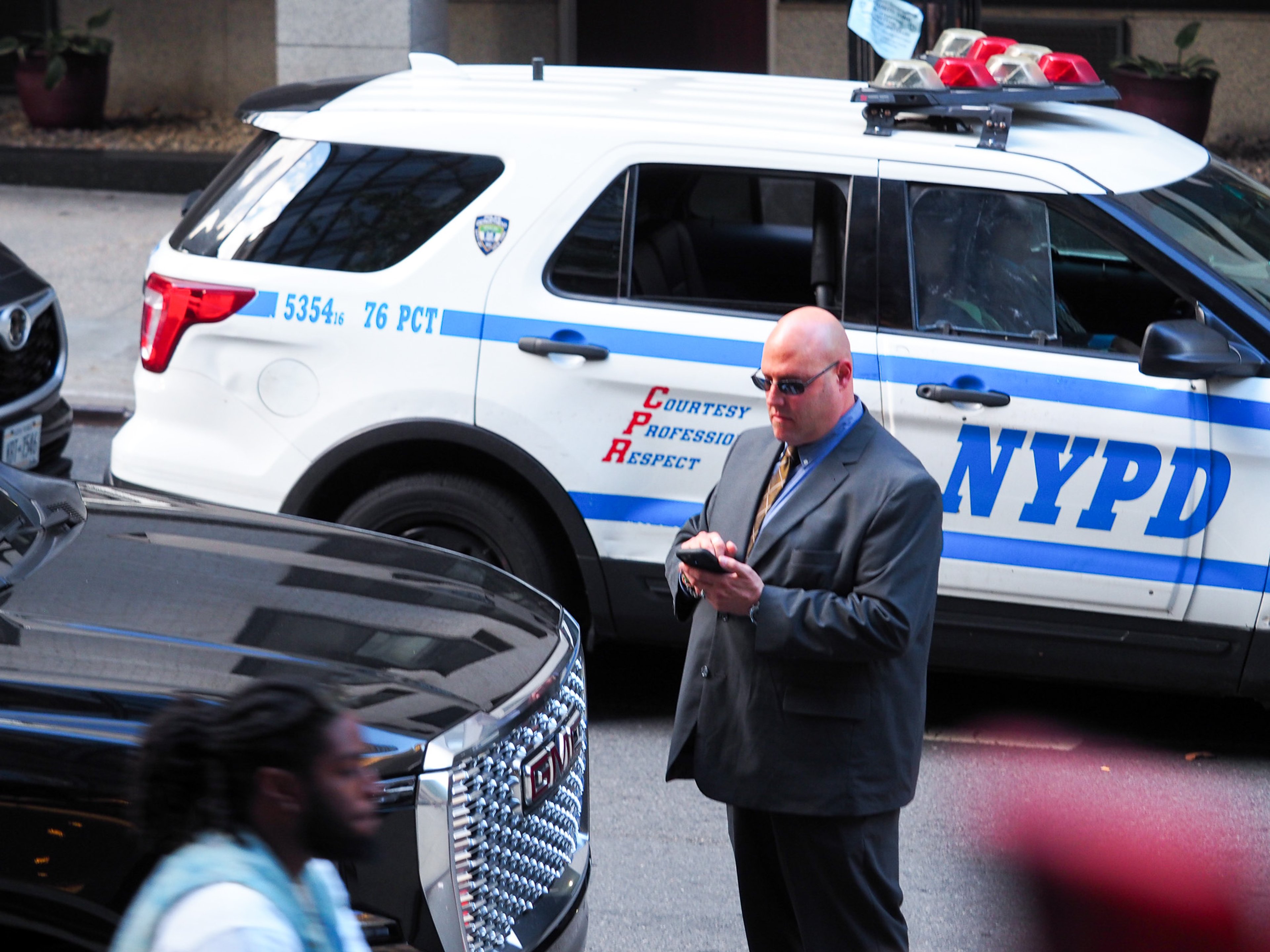 A bald man in a dark suit, sunglasses, and gold tie checks his phone in front of an NYPD SUV reading '5354, 76 PCT, Courtesy, Professionalism, Respect,' with a black motorcade vehicle in the foreground and a pedestrian passing.
