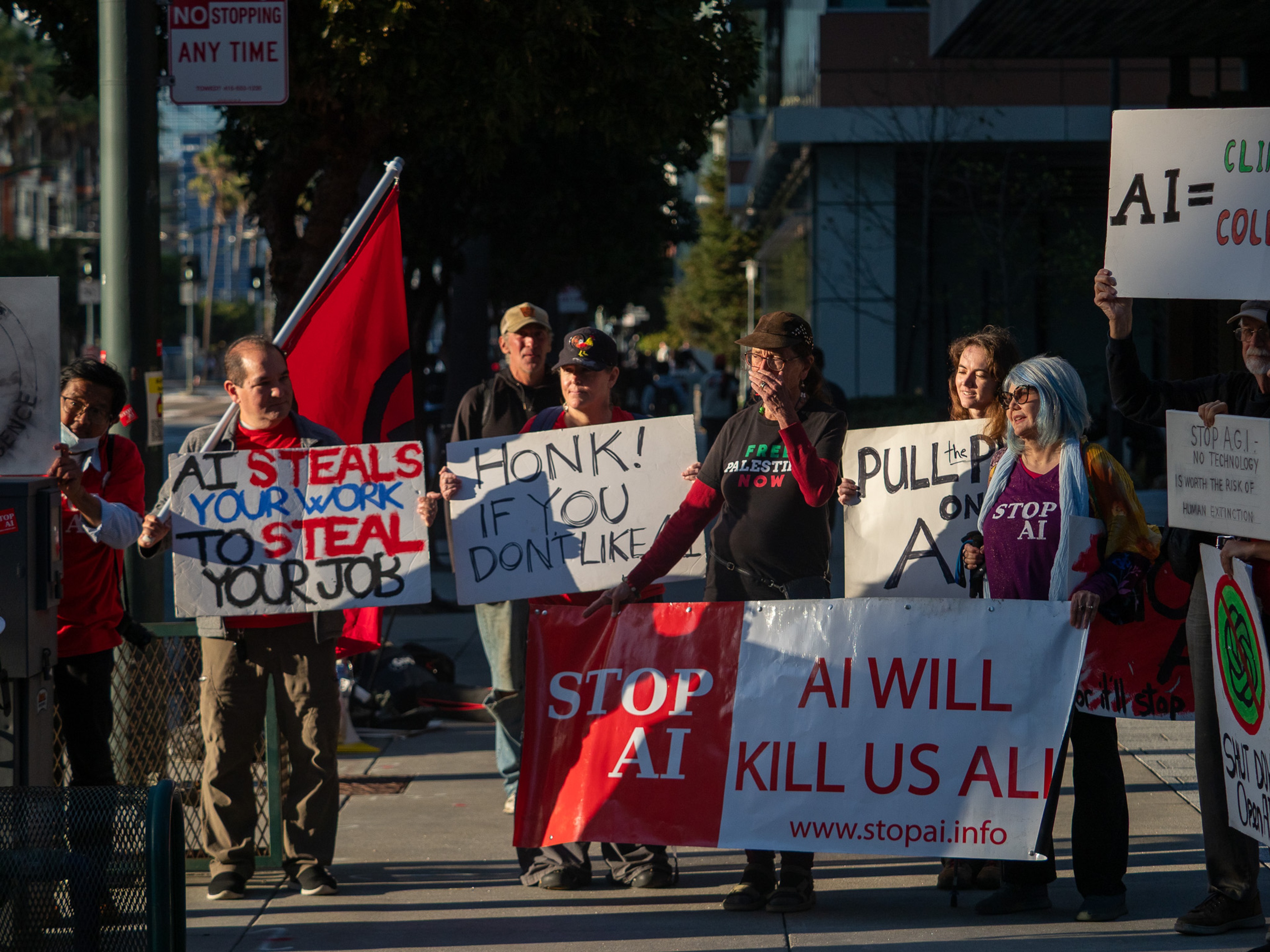 A line of protesters on a downtown sidewalk holds signs reading 'Stop AI', 'AI Will Kill Us All', 'AI Steals Your Work to Steal Your Job', and 'Honk if you don't like AI', backed by a red flag.