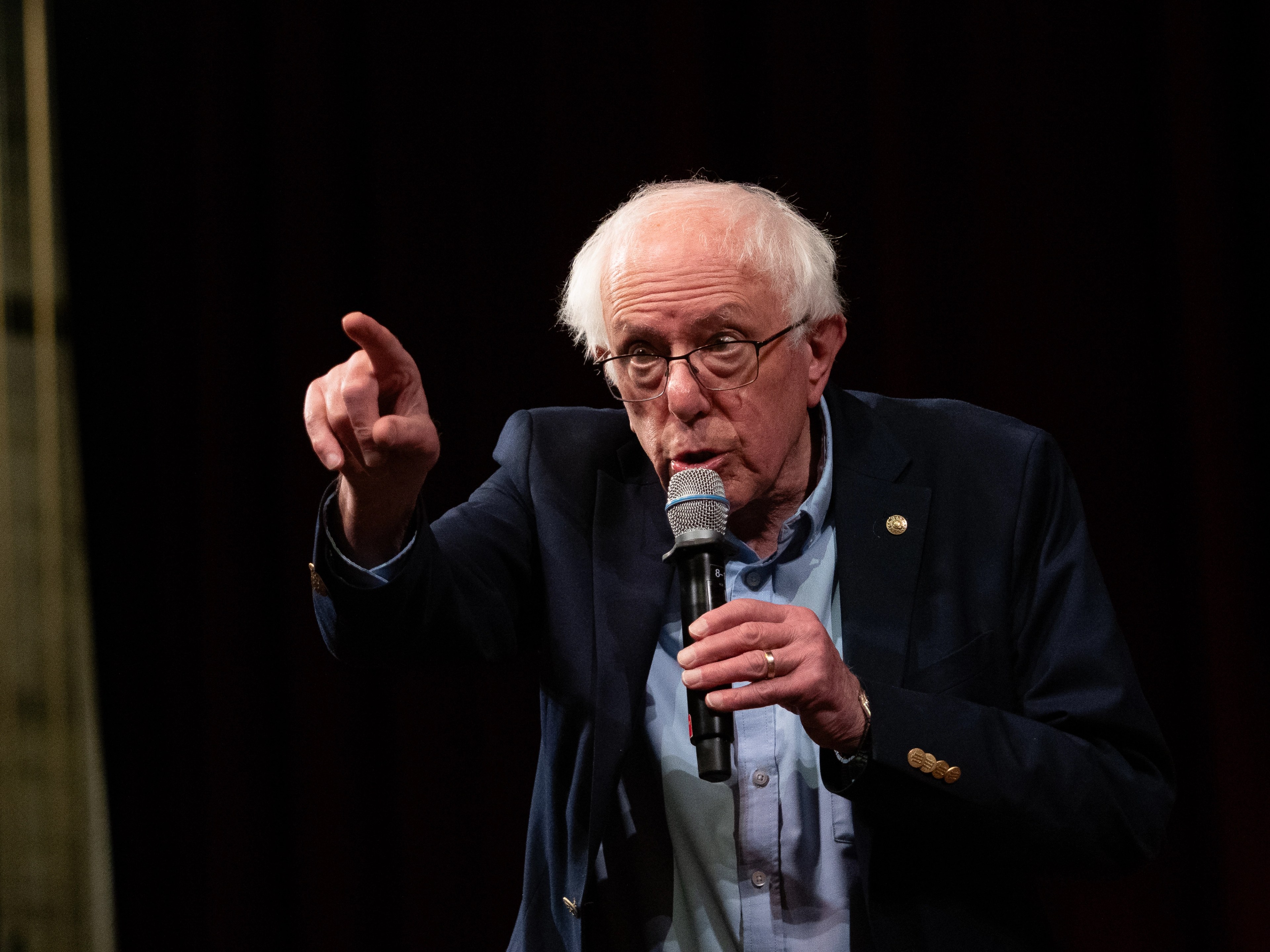 Tight portrait of Bernie Sanders speaking, hand raised mid-gesture, holding a microphone close to his face, partial American flag visible to the right against a dark stage background.