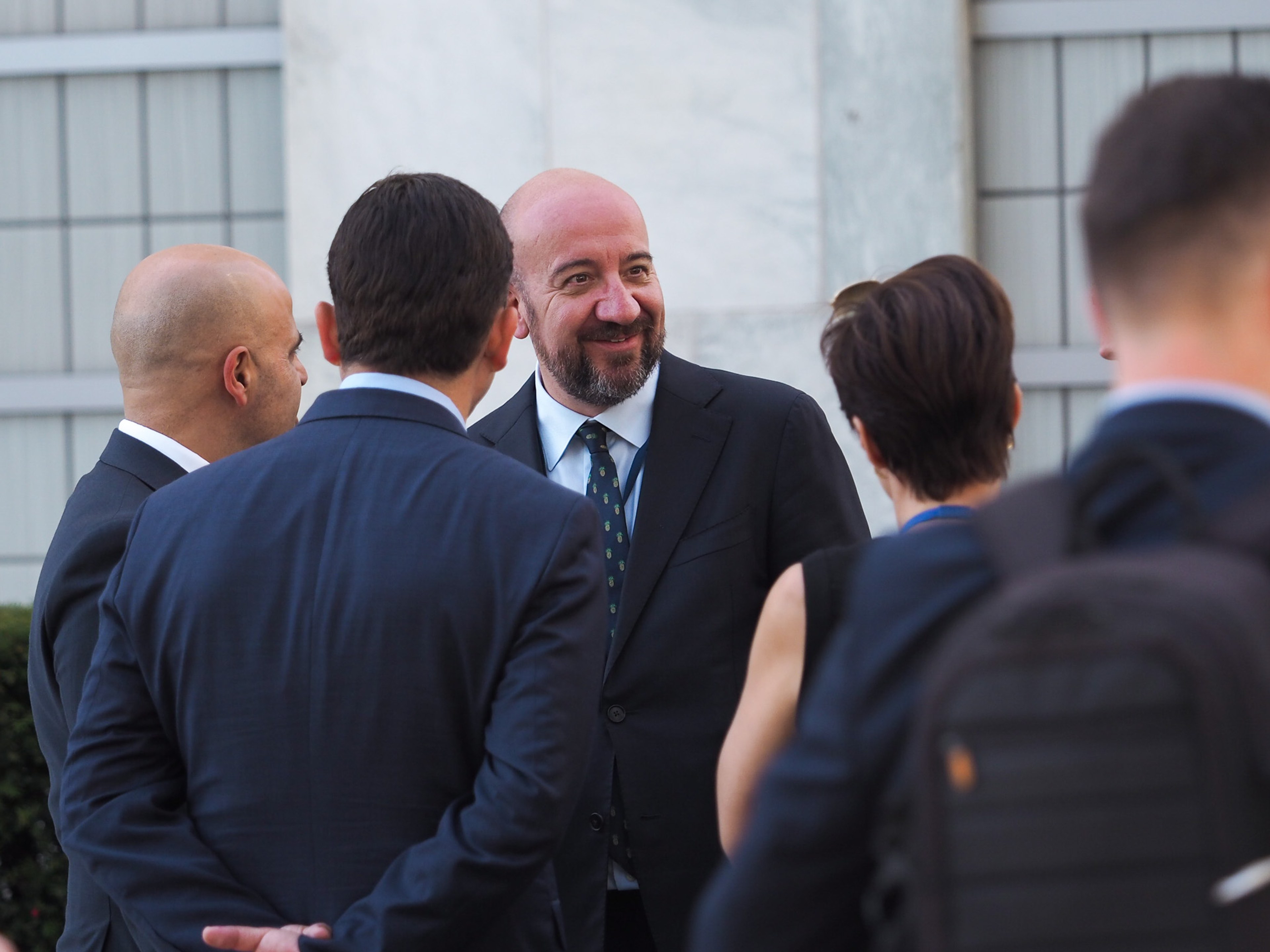 Charles Michel, salt-and-pepper beard, smiles warmly mid-conversation outside a marble UN building, surrounded by aides — backs of heads, a woman with short dark hair, a young man with a backpack.