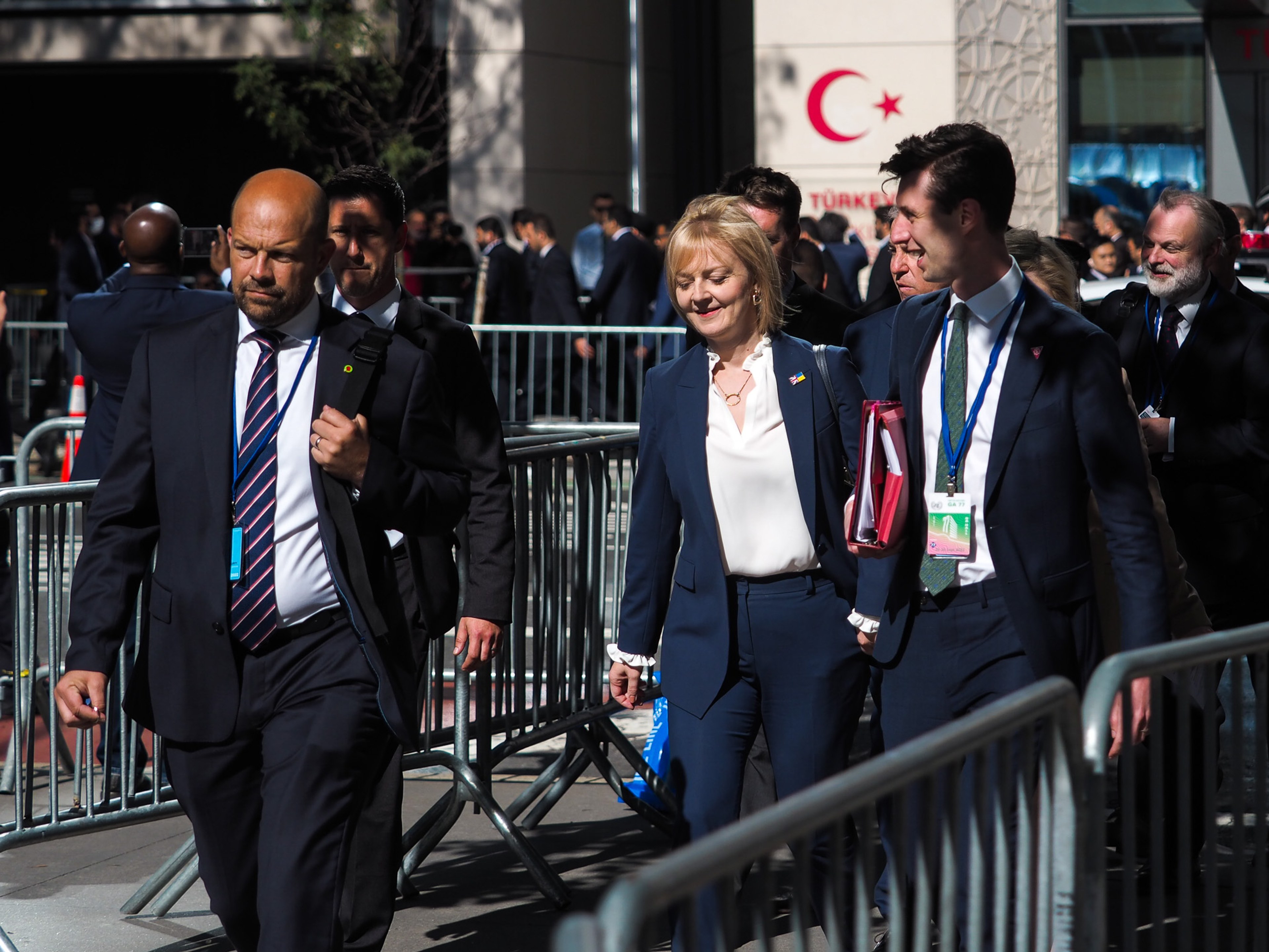 British Prime Minister Liz Truss in a navy suit walks through metal crowd barriers flanked by a bald security agent and a young aide holding red folders, the Turkish pavilion banner reading 'TÜRKİYE' visible behind.