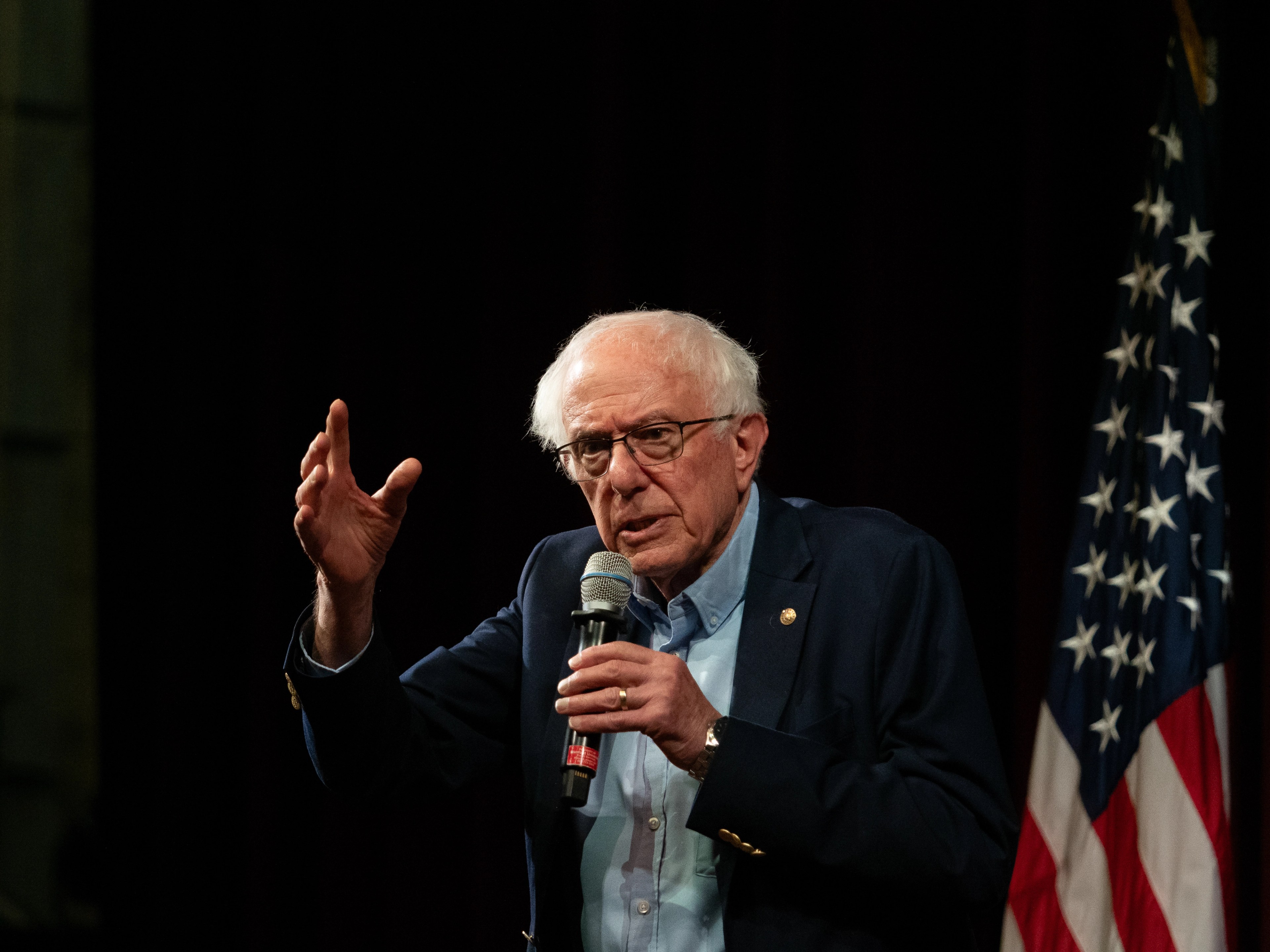 Bernie Sanders behind a wooden podium with one hand raised palm-up mid-gesture, the other resting on the podium, the red microphone-icon logo visible on the front.