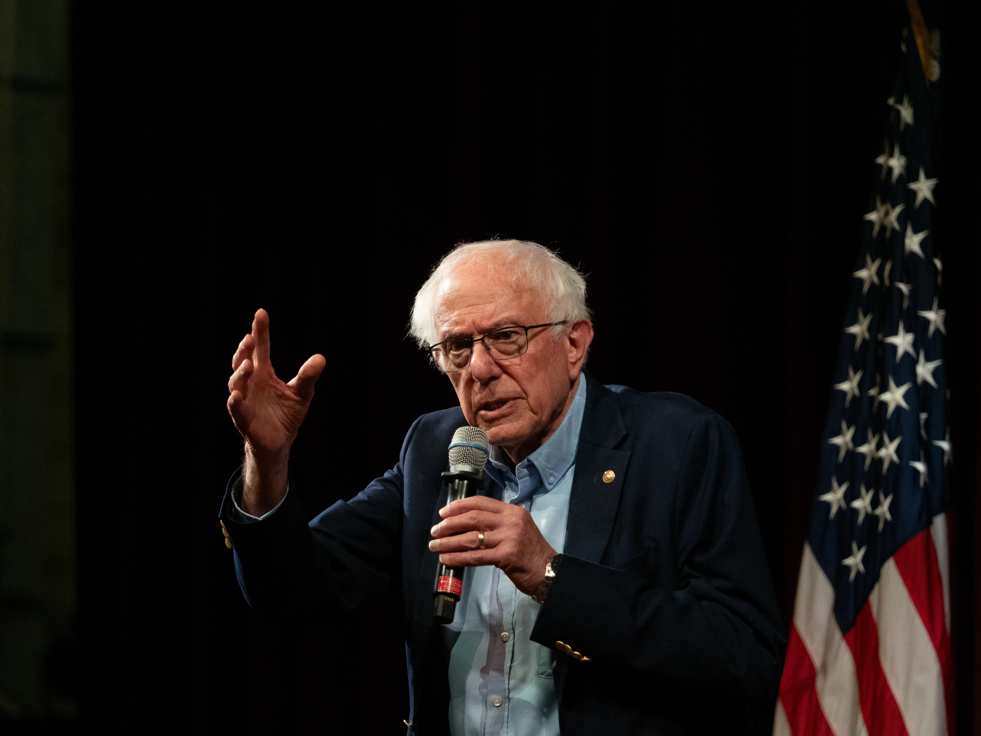 Bernie Sanders behind a wooden podium with one hand raised palm-up mid-gesture, the other resting on the podium, the red microphone-icon logo visible on the front.