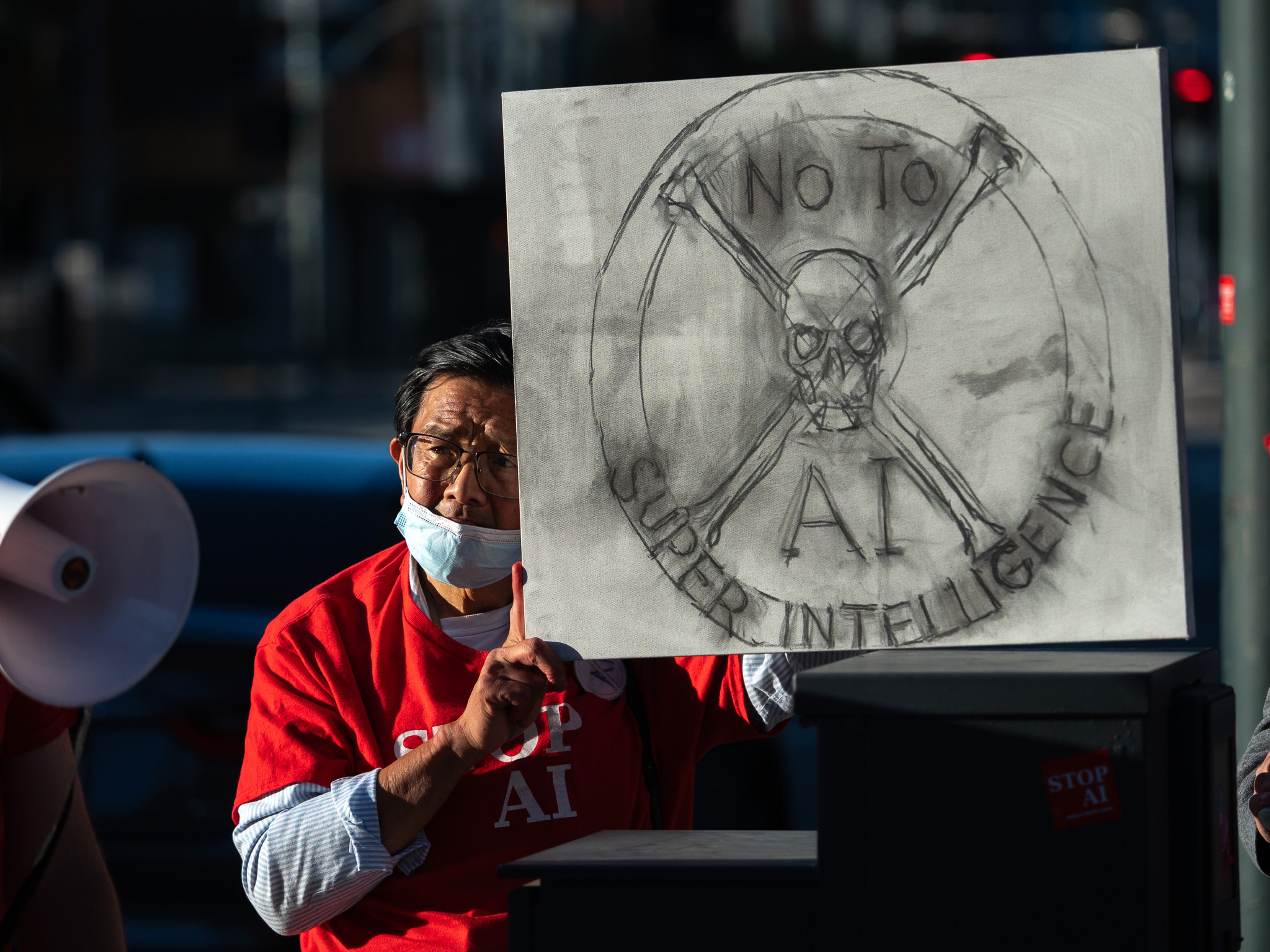 A protester in a red Stop AI shirt and surgical mask holds up a hand-drawn placard depicting a skull inside a radiation symbol, reading 'No to Super Intelligence'.