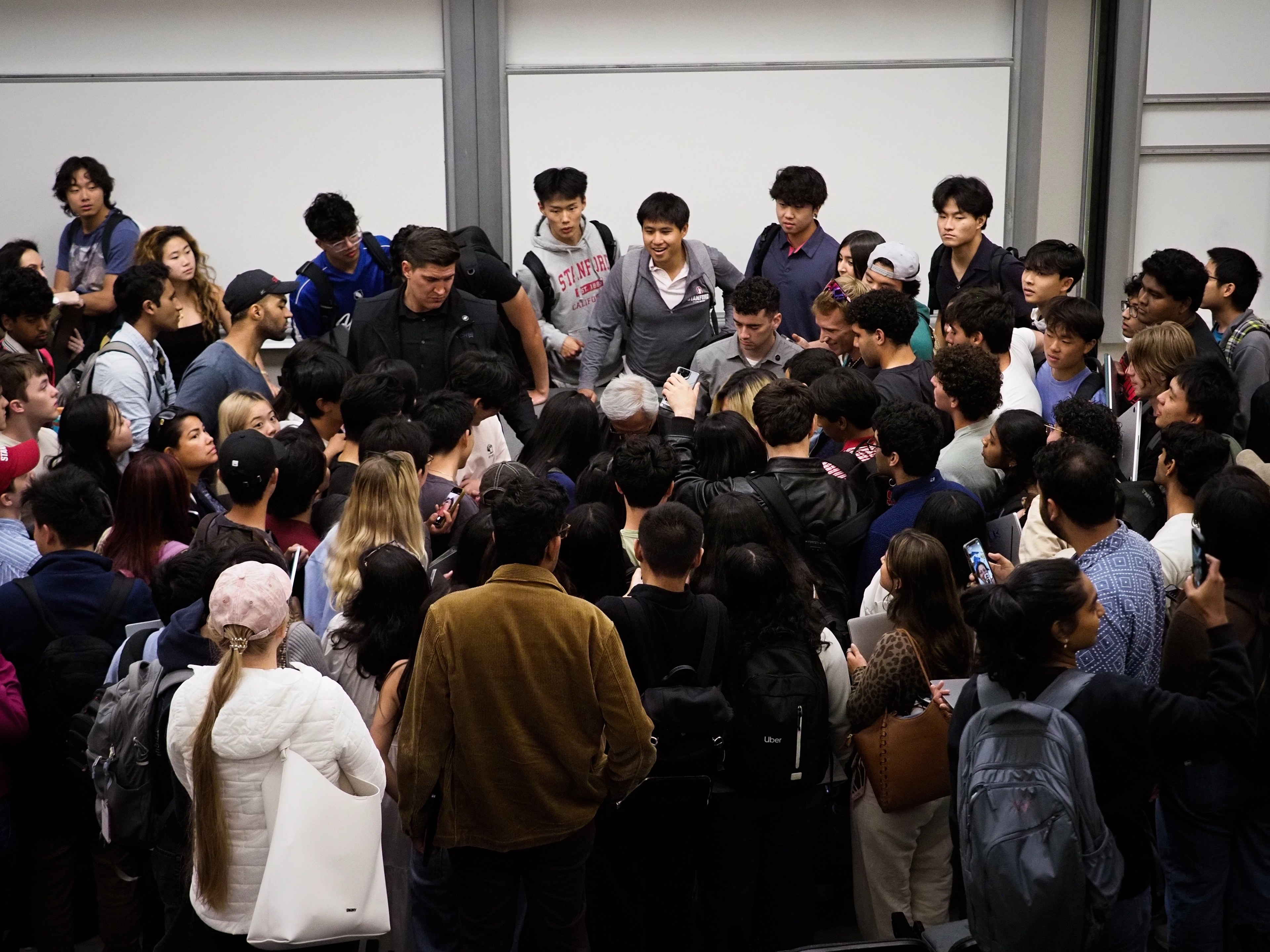 Wide overhead view of dozens of Stanford students packed together in a hallway, Jensen Huang barely visible at the center of the swarm, several students wearing Stanford sweatshirts in the upper portion of the crowd.