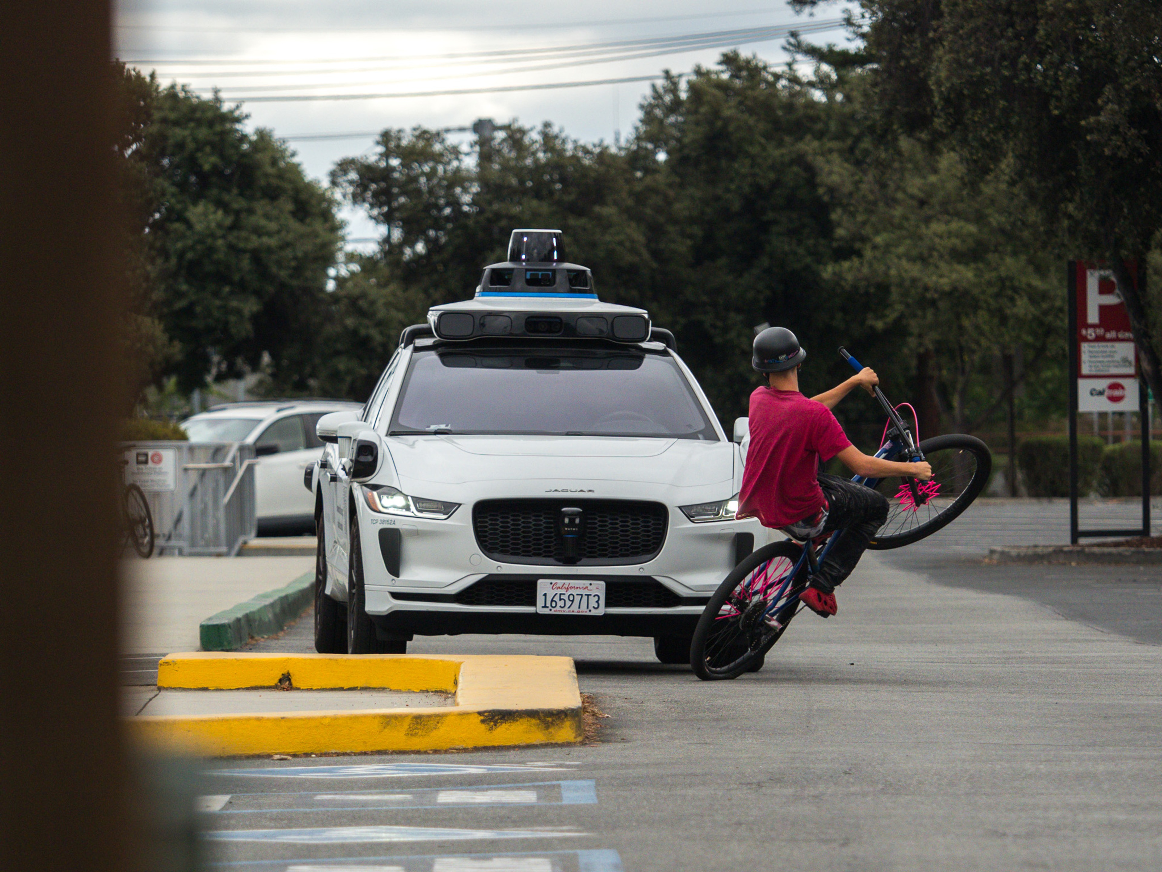 A young man on a BMX bicycle holds a wheelie in front of a white Waymo robotaxi as the front wheel begins to come down.