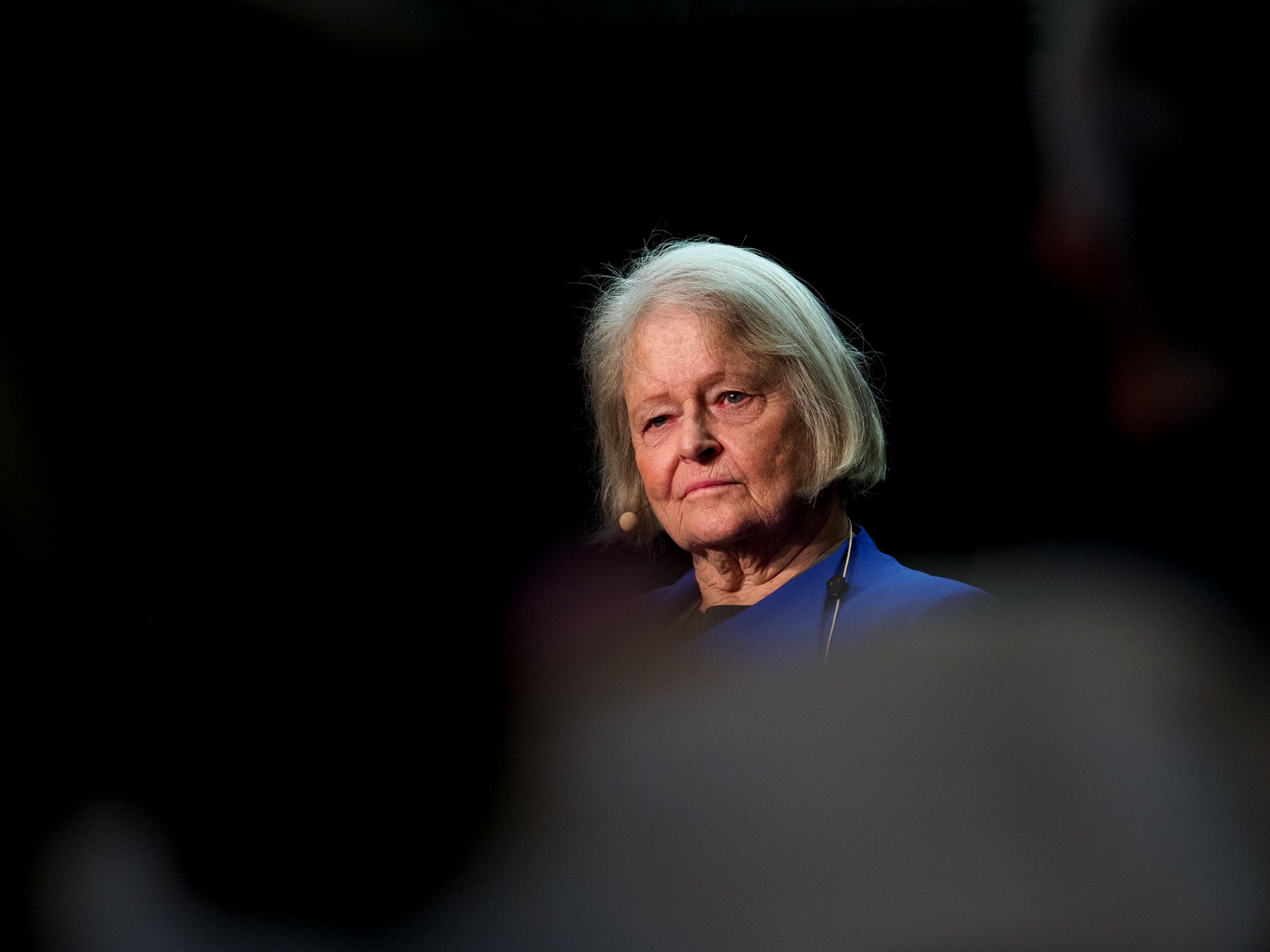 Tight portrait of Gro Harlem Brundtland in a blue jacket with a thin headset microphone, looking forward against a deep black background, the out-of-focus shoulders of audience members framing the lower corners of the frame.