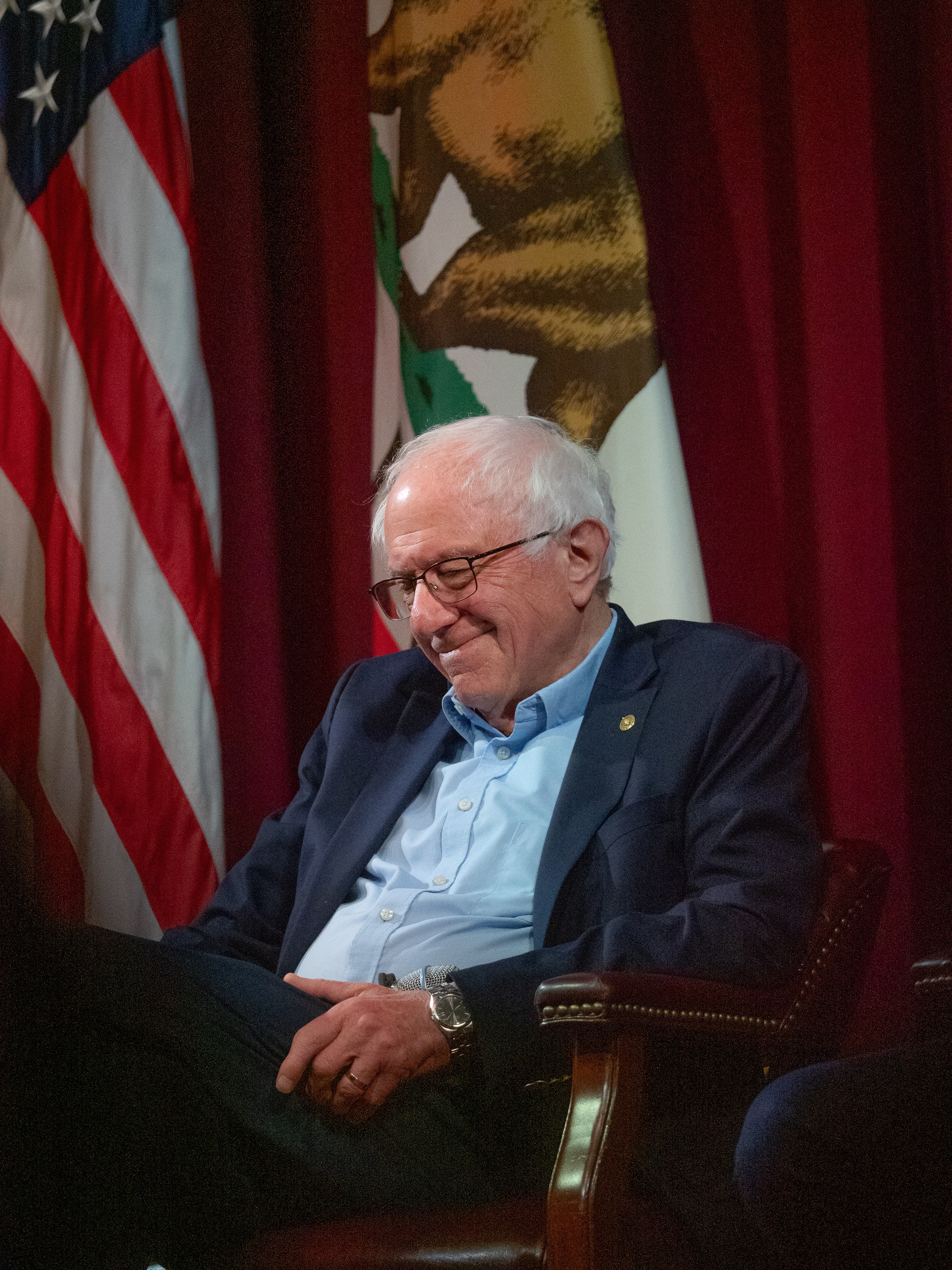 Bernie Sanders seated in a leather chair, smiling and looking down, framed by an American flag and California state flag against a deep red curtain.