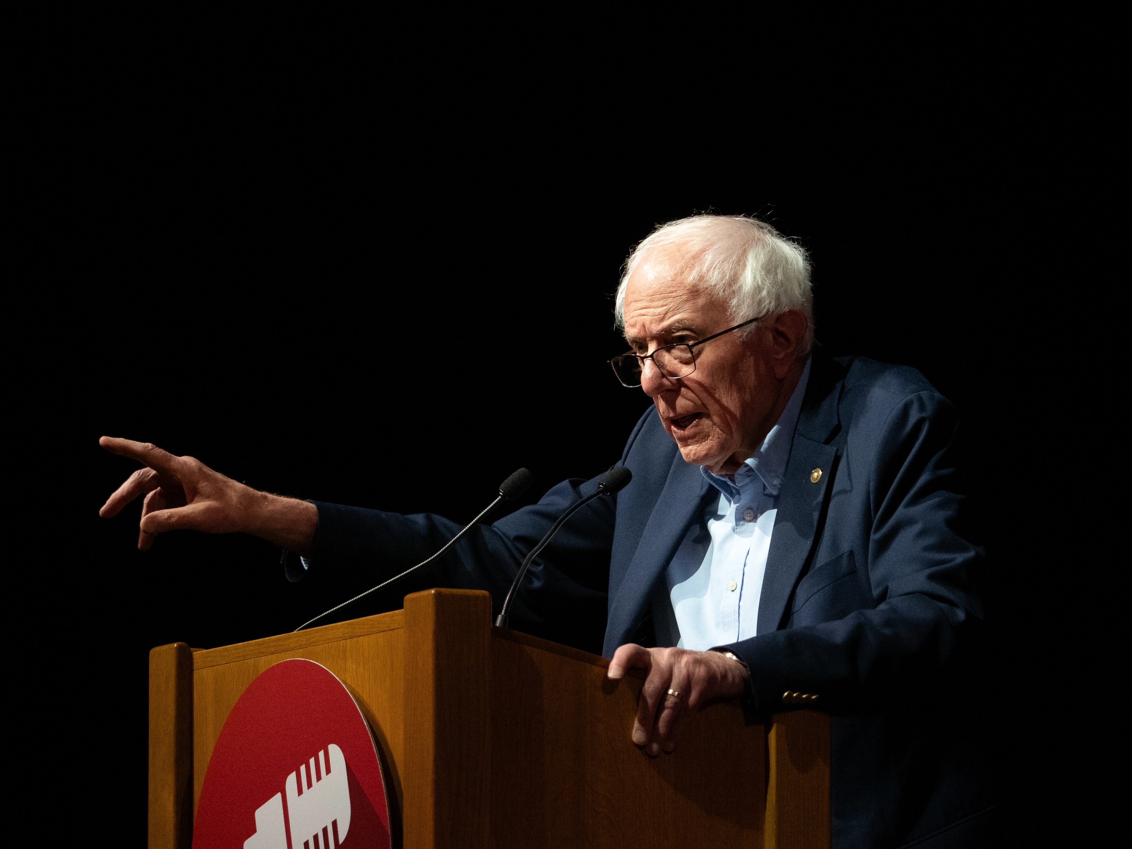 Senator Bernie Sanders at a wooden podium, glasses on the tip of his nose, finger pointed toward the audience as he speaks into a microphone, with a red microphone-icon logo visible on the front of the podium.